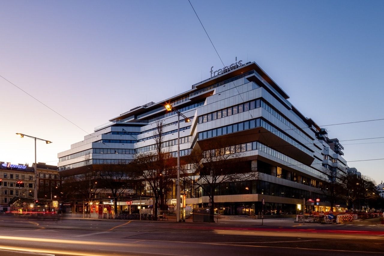 Modernes Bürogebäude "Francis" in der Dämmerung mit beleuchteten Fenstern und Straßenverkehr.