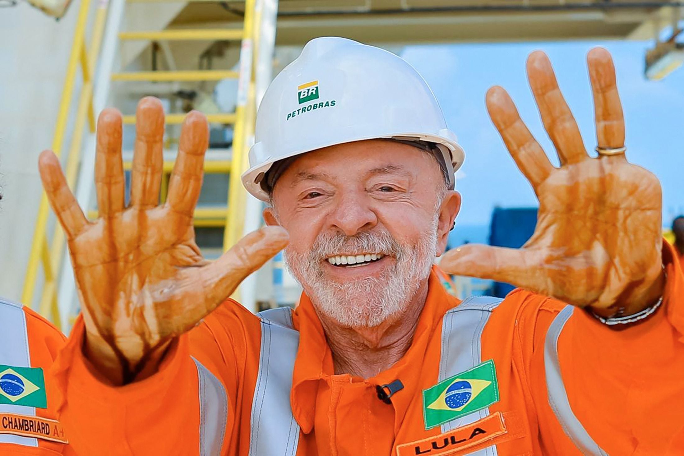 Ein Mann im orangefarbenen Overall zeigt seine ölverschmierten Hände und lächelt in die Kamera.
