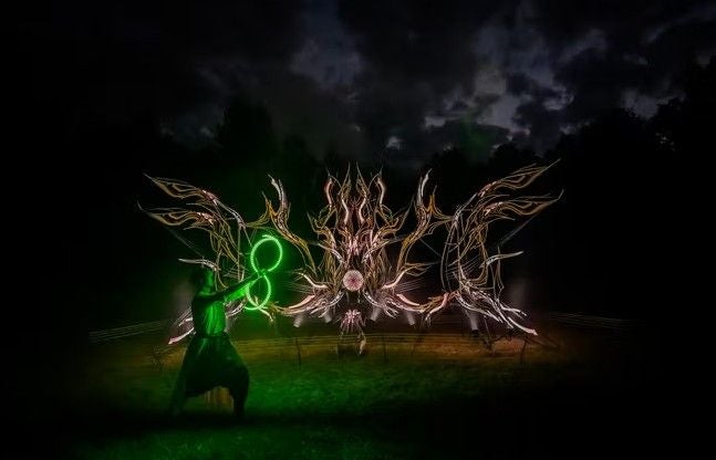 Person mit grünen Lichteffekten vor einer kunstvollen, leuchtenden Installation im Dunkeln.