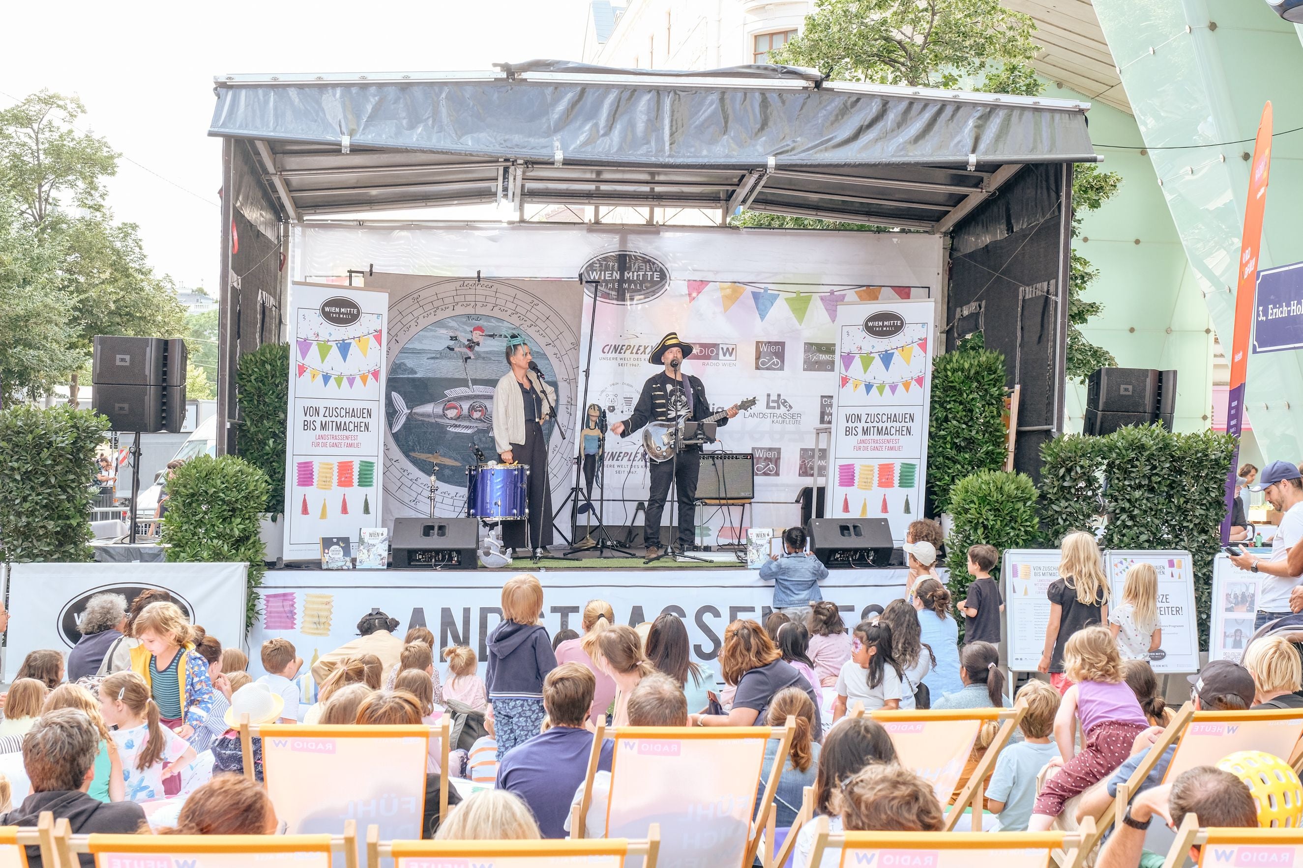 Zwei Musiker treten auf einer Bühne beim Landstraßenfest vor einem sitzenden Kinderpublikum auf.