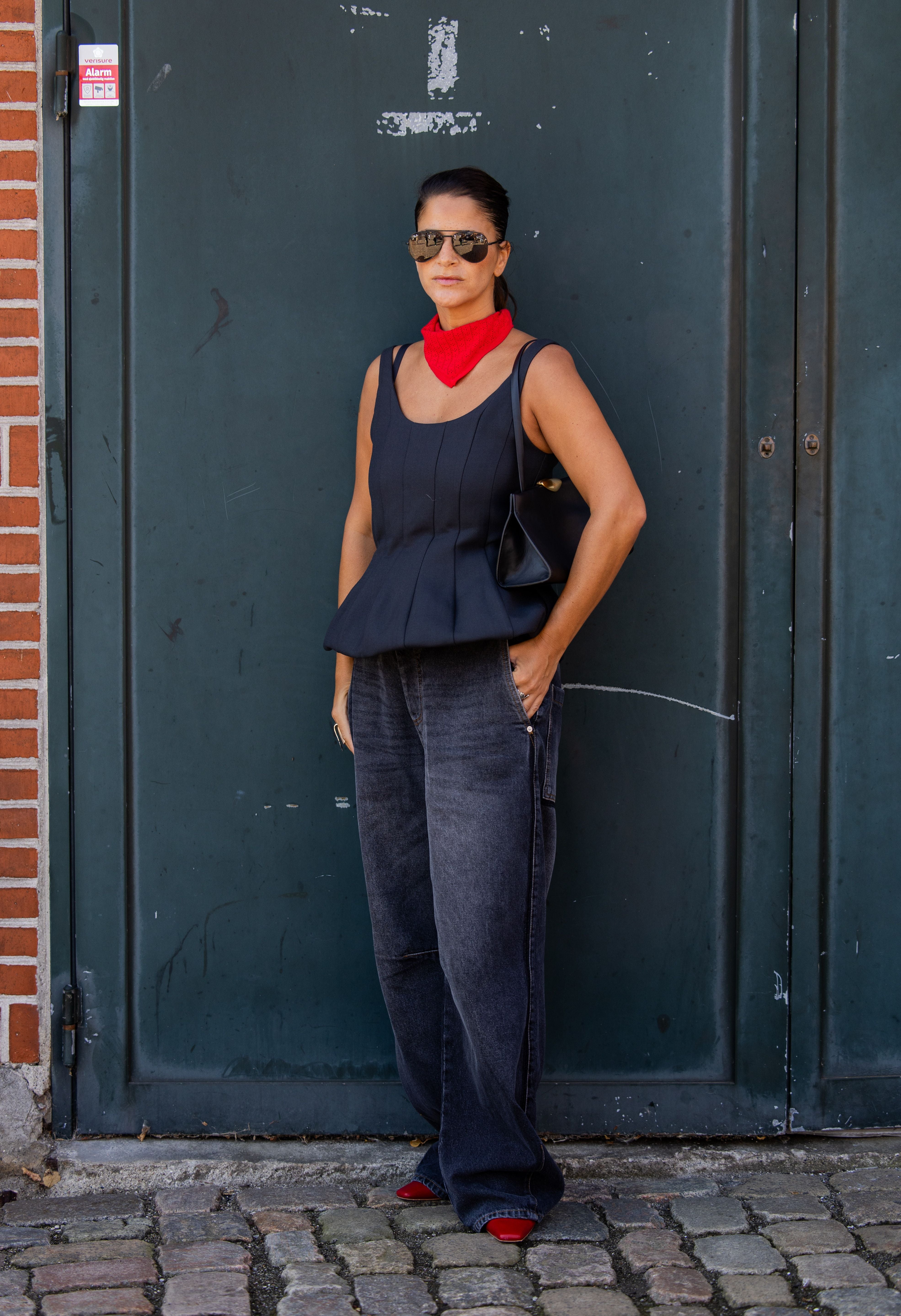 Frau mit rotem Halstuch, grauem Top, dunkler Jeans und Sonnenbrille steht vor grünem Tor.