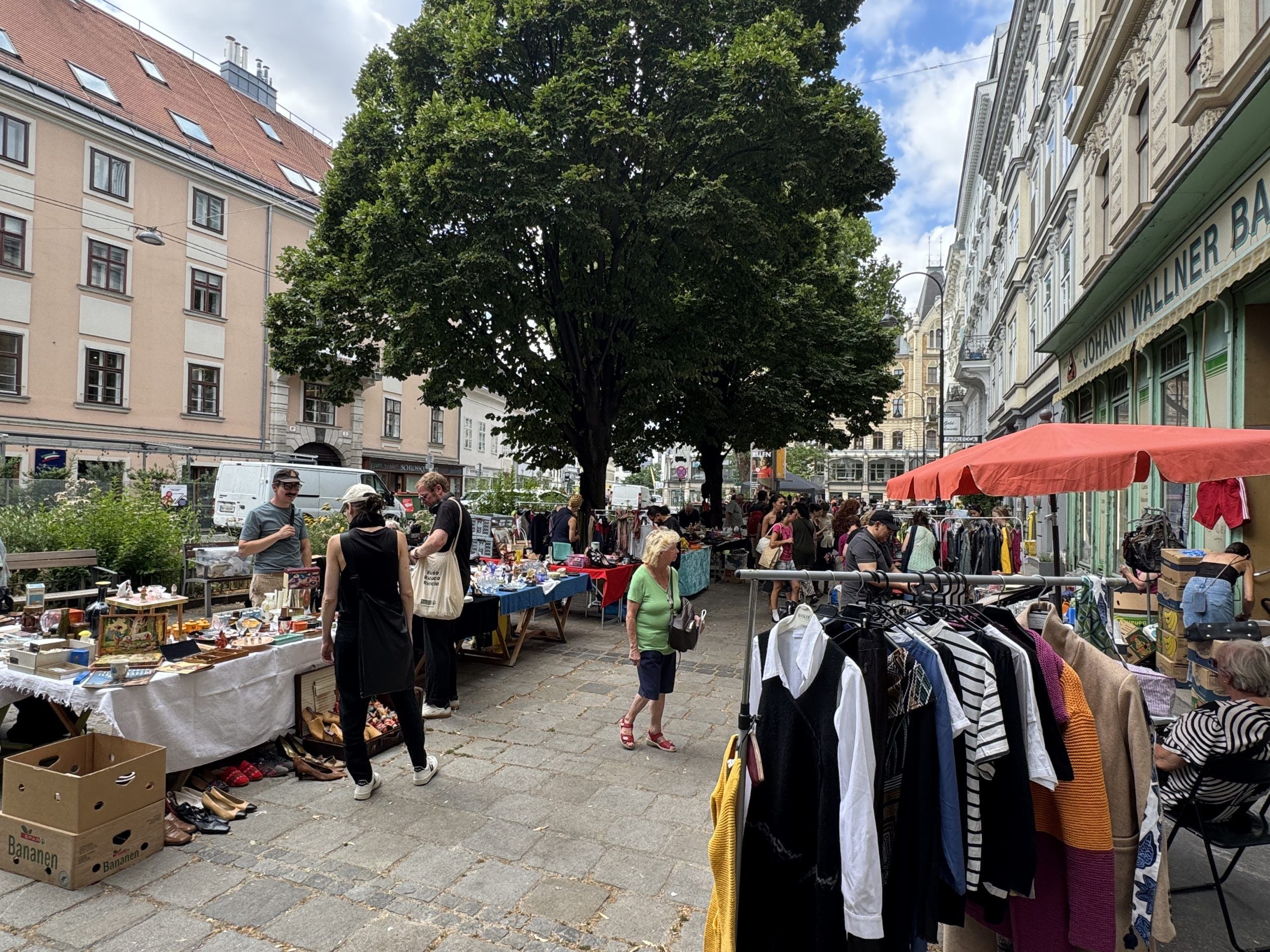 Flohmarkt mit Ständen unter Bäumen, Kleidung und verschiedene Waren werden angeboten.