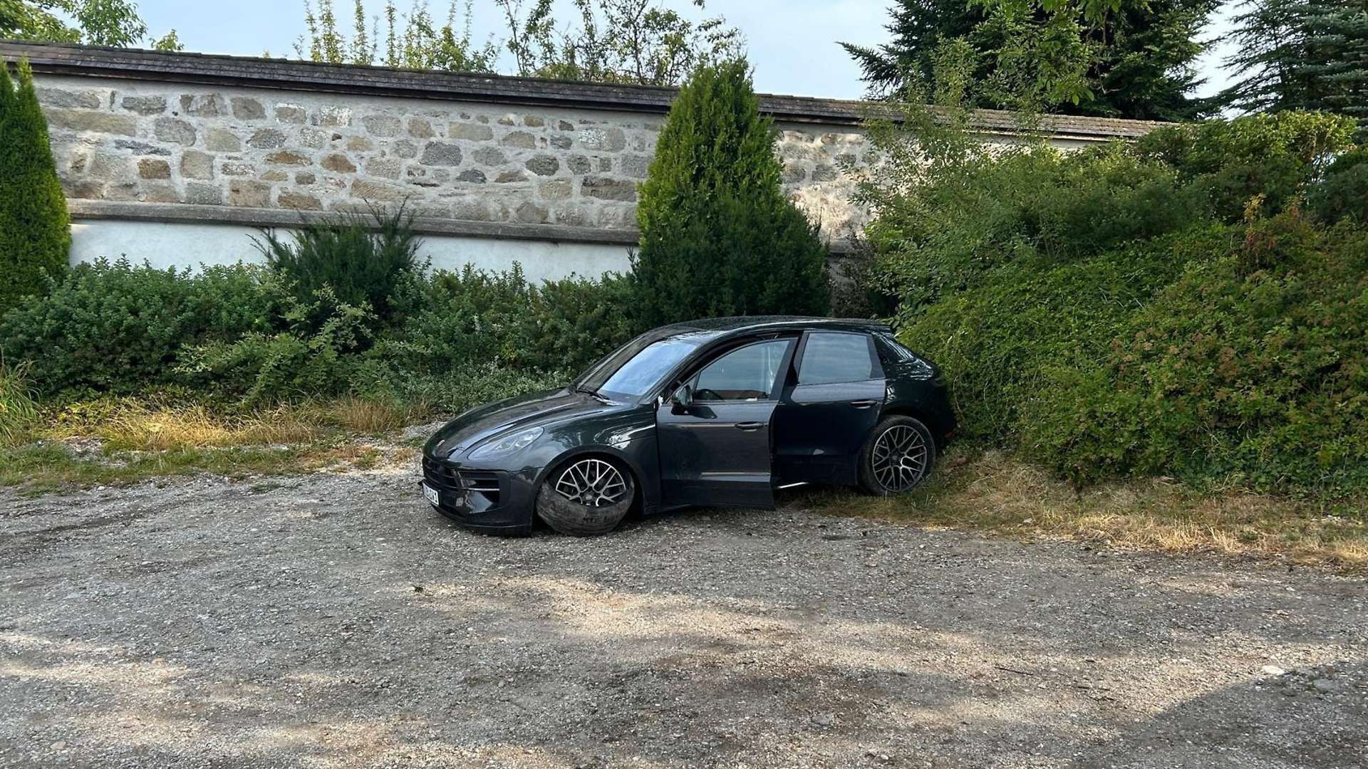 Schwarzer Porsche mit offener Tür steht beschädigt am Straßenrand vor einer Steinmauer und Büschen.