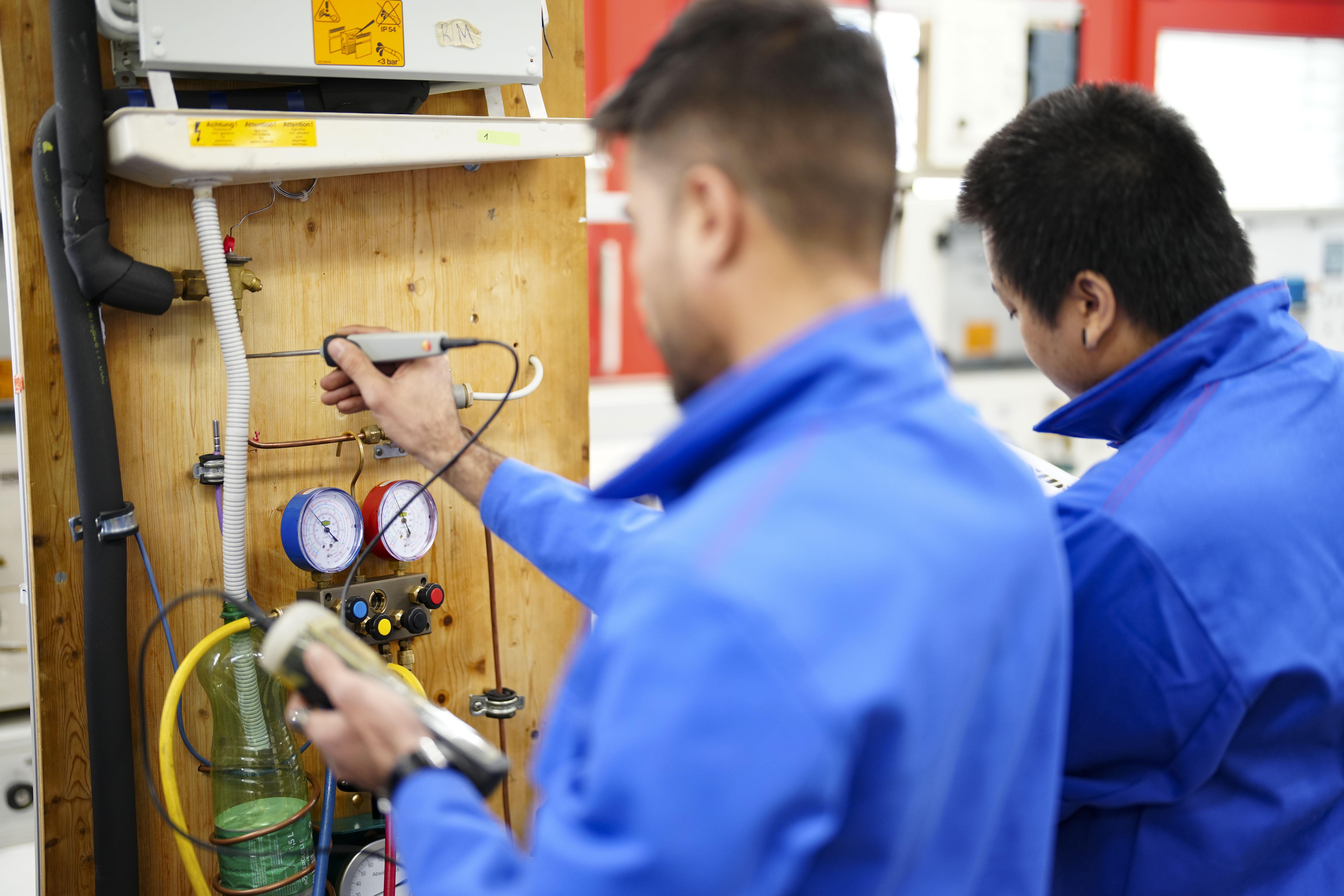 Zwei junge Männer in blauer Arbeitskleidung arbeiten an einer technischen Anlage.