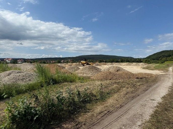 Schotterhügel und Bagger auf einer Baustelle, im Hintergrund Hügel und Häuser.