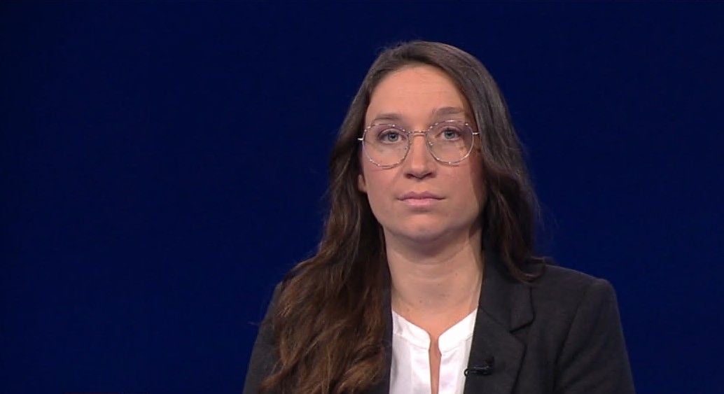 Eine Frau mit Brille und langen braunen Haaren vor blauem Hintergrund.