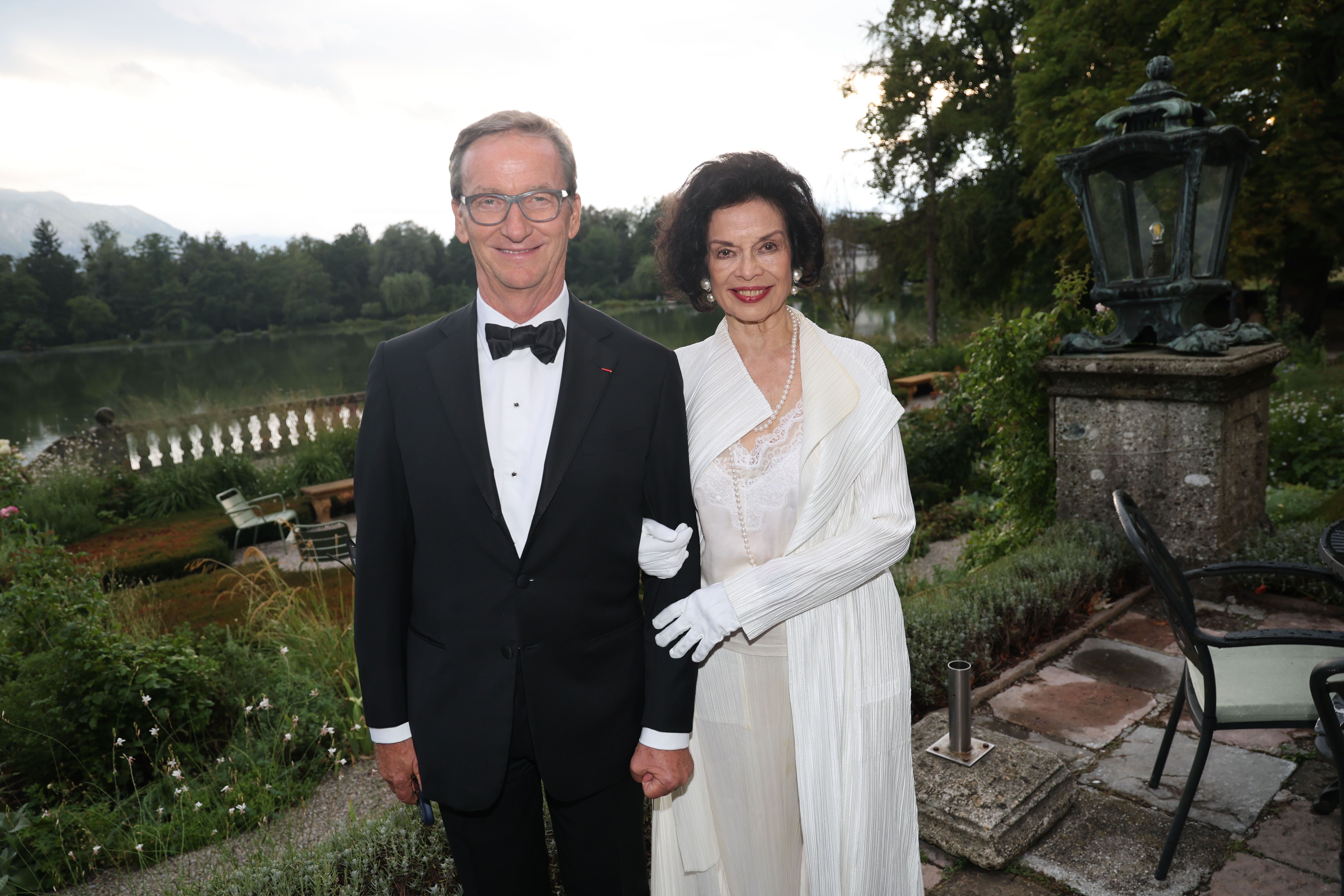 Ein Mann im Smoking und eine Frau in weißem Abendkleid posieren im Garten vor Schloss Leopoldskron.