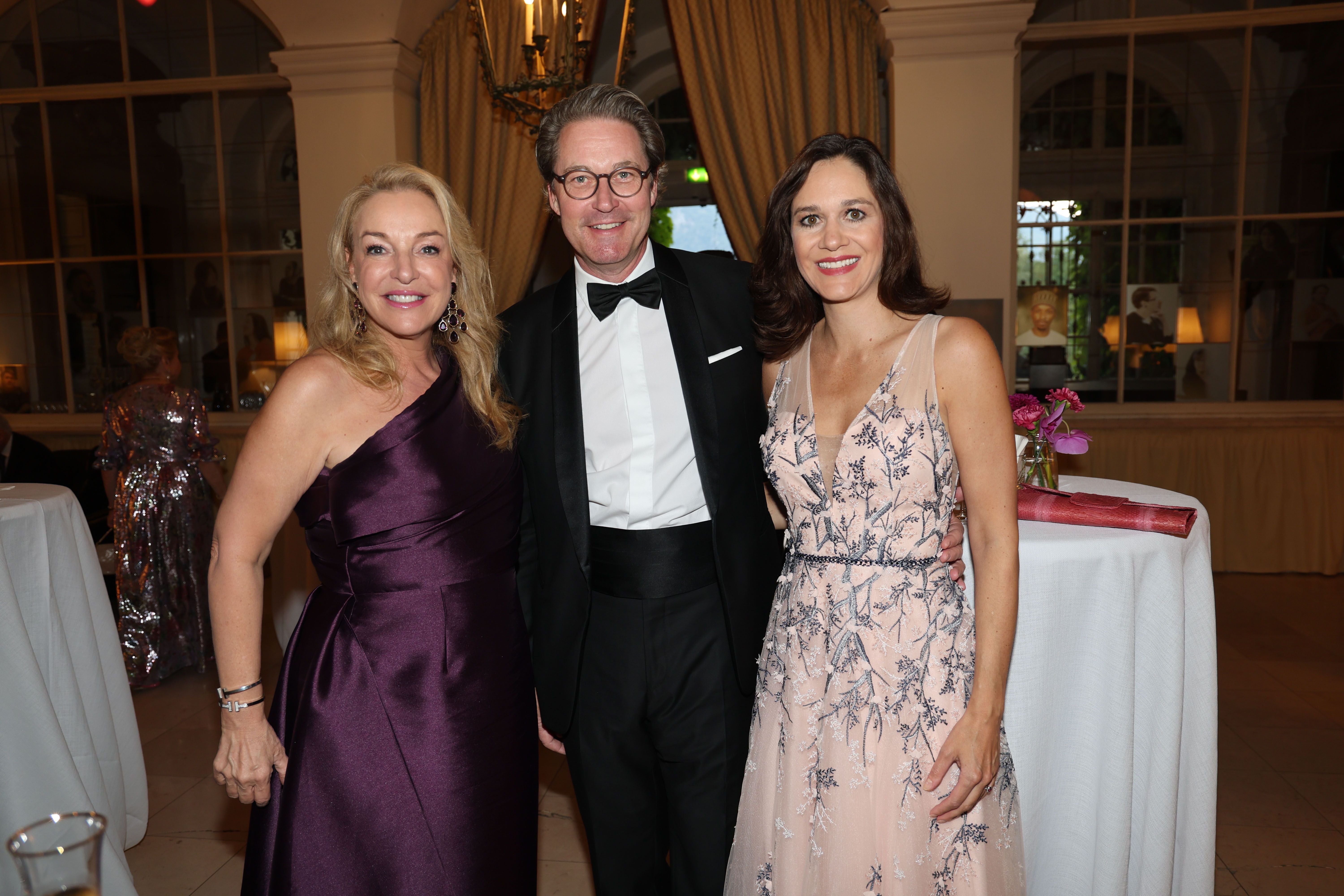 Drei elegant gekleidete Personen posieren bei einer Gala im festlich dekorierten Saal.