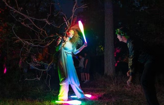 Eine Person in Kostüm jongliert bei Nacht mit leuchtenden Keulen im Wald.
