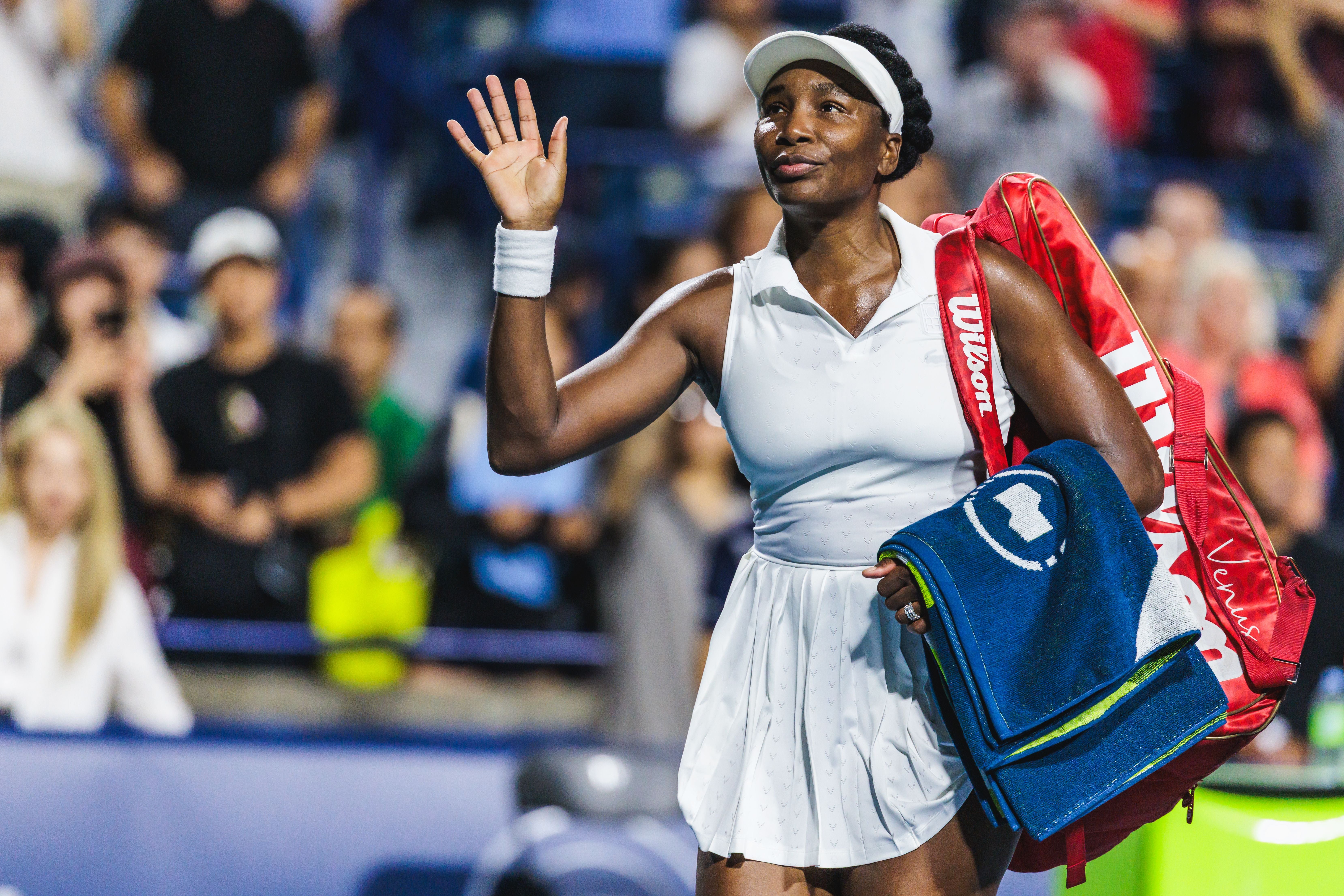 Venus Williams winkt in weißem Tennis-Outfit mit erhobener Hand den Fans zu, während sie ihre Tasche und ein Handtuch über der Schulter trägt.