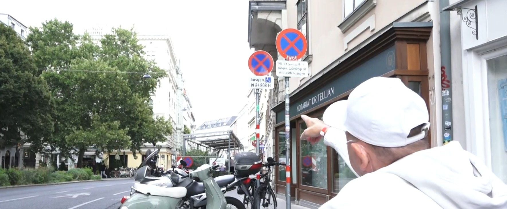 Person mit weißer Kappe deutet auf Halteverbotsschilder in einer Wiener Straße mit Mopeds.