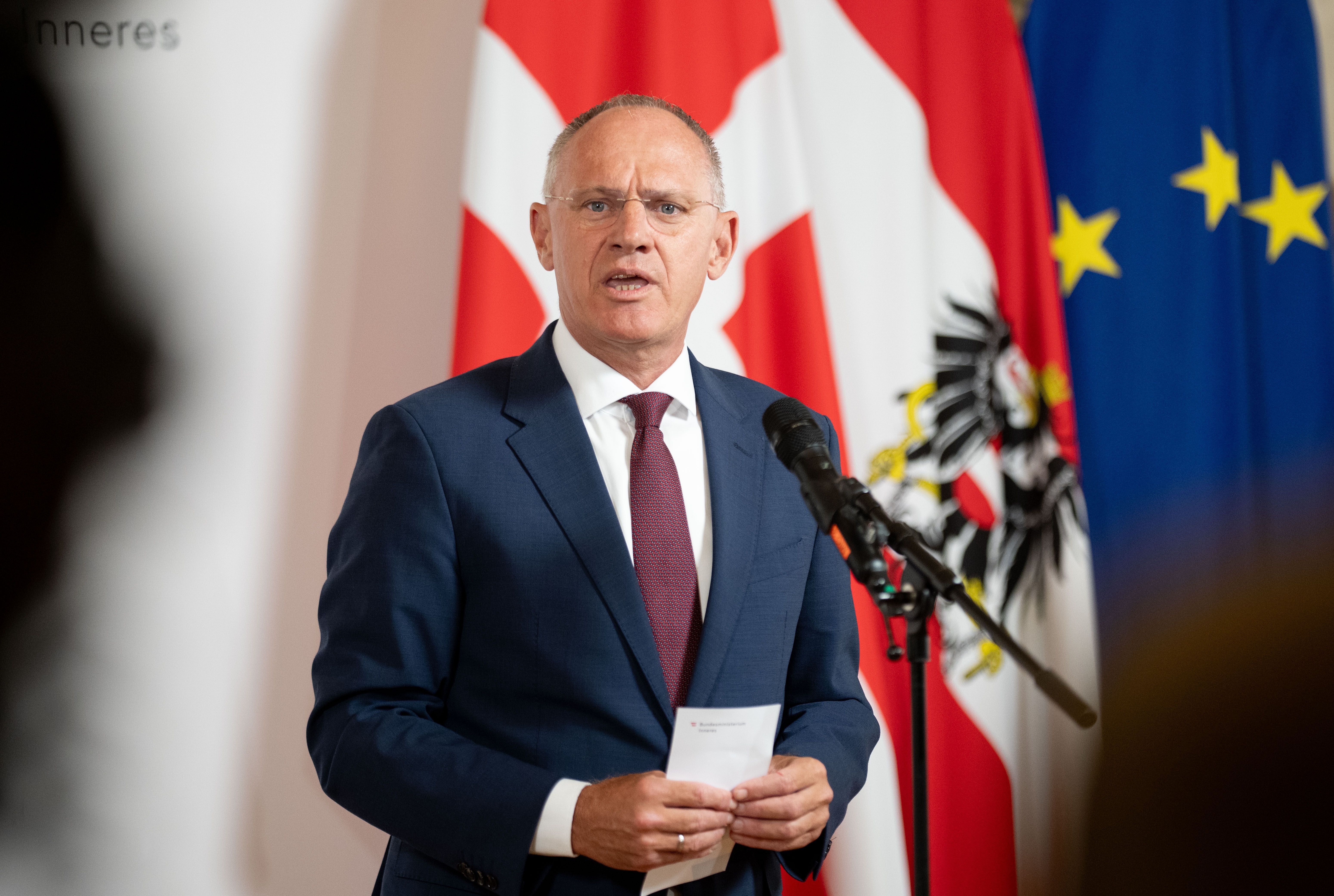 Ein Mann im Anzug spricht bei einer Pressekonferenz vor österreichischer und EU-Flagge.