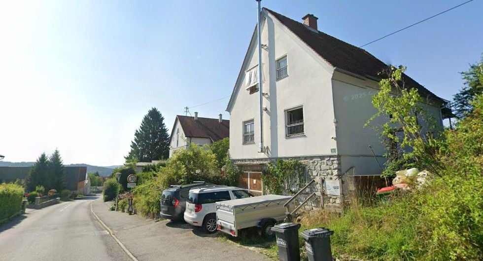 Ein weißes Wohnhaus an einer Straße mit Autos und Mülltonnen im Vordergrund.