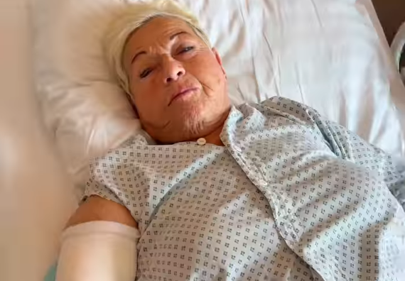 Ältere Frau liegt mit verbundenem Arm im Krankenhausbett und trägt ein Krankenhaushemd.