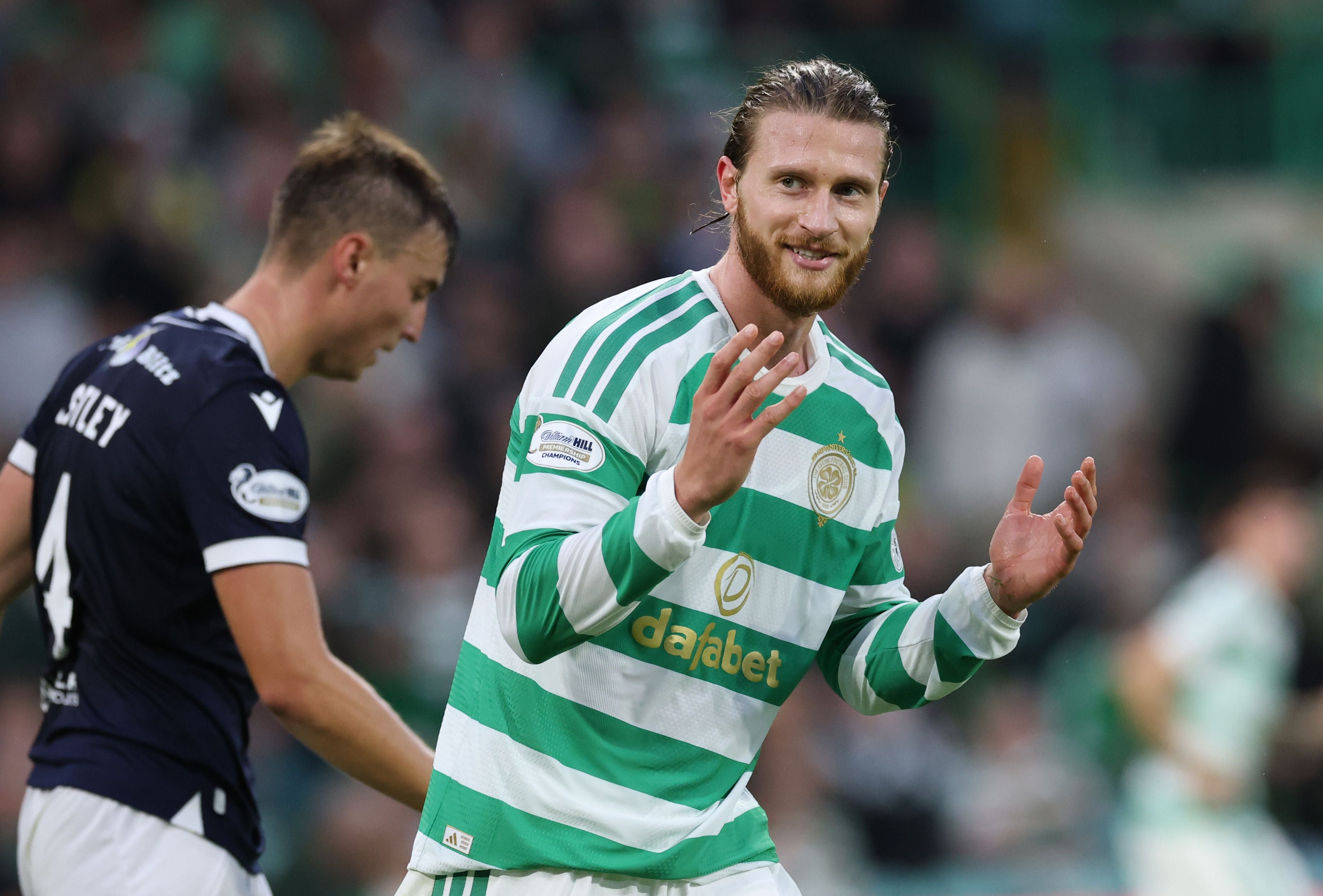 Kasper Høgh im grün-weiß gestreiften Celtic Glasgow Trikot blickt mit gehobenen Händen lächelnd zur Seite, während im Hintergrund ein Gegenspieler in dunklem Trikot vorbeiläuft.