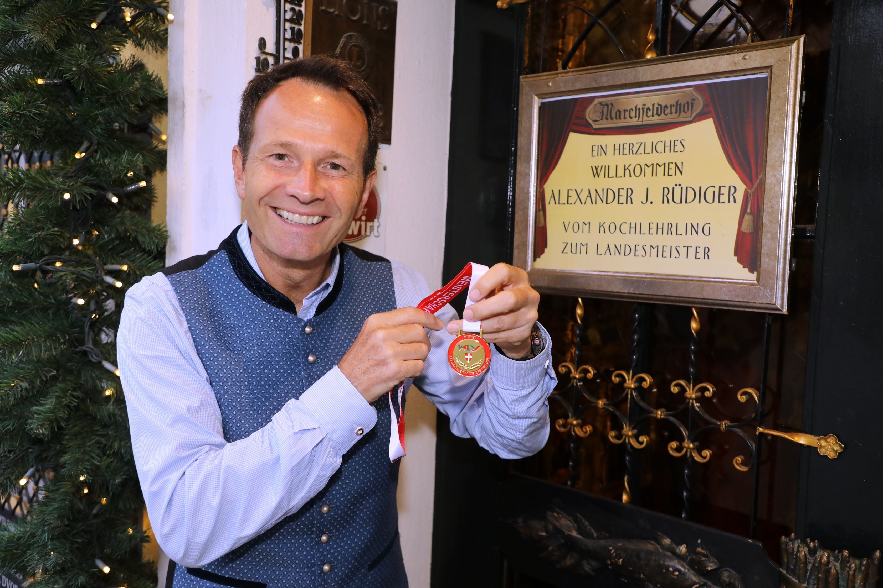 Ein Mann hält lächelnd eine Medaille vor einem Begrüßungsschild im festlich dekorierten Raum.