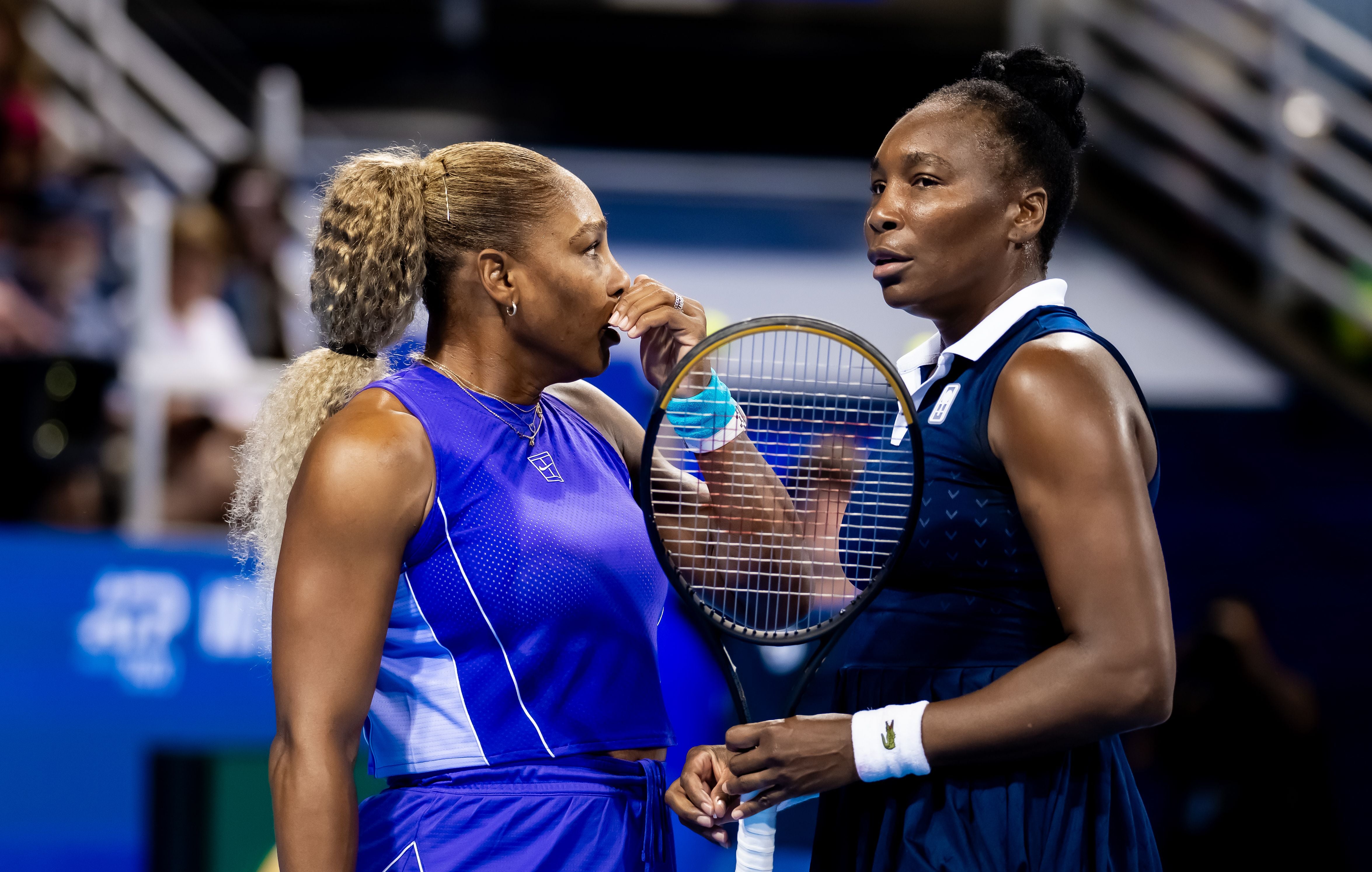 Serena und Venus Williams stehen nebeneinander auf einem Tennisplatz, schauen sich an und besprechen sich auf dem Court.