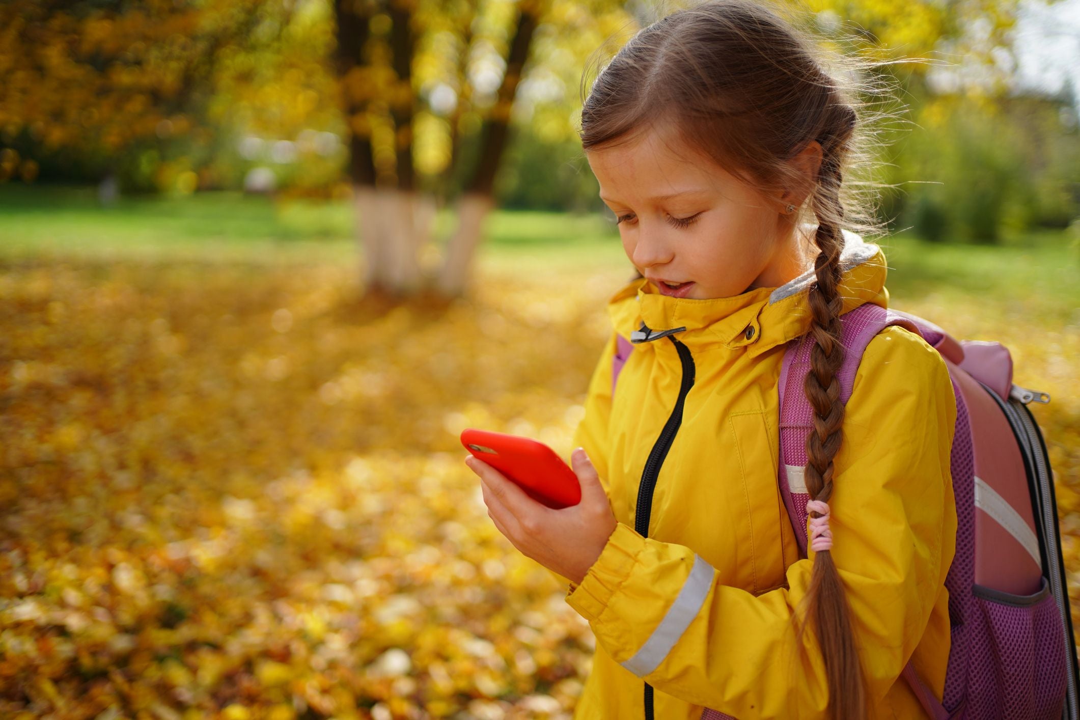Schulmädchen im gelben Mantel macht einen Videoanruf mit dem Smartphone im Herbstpark.