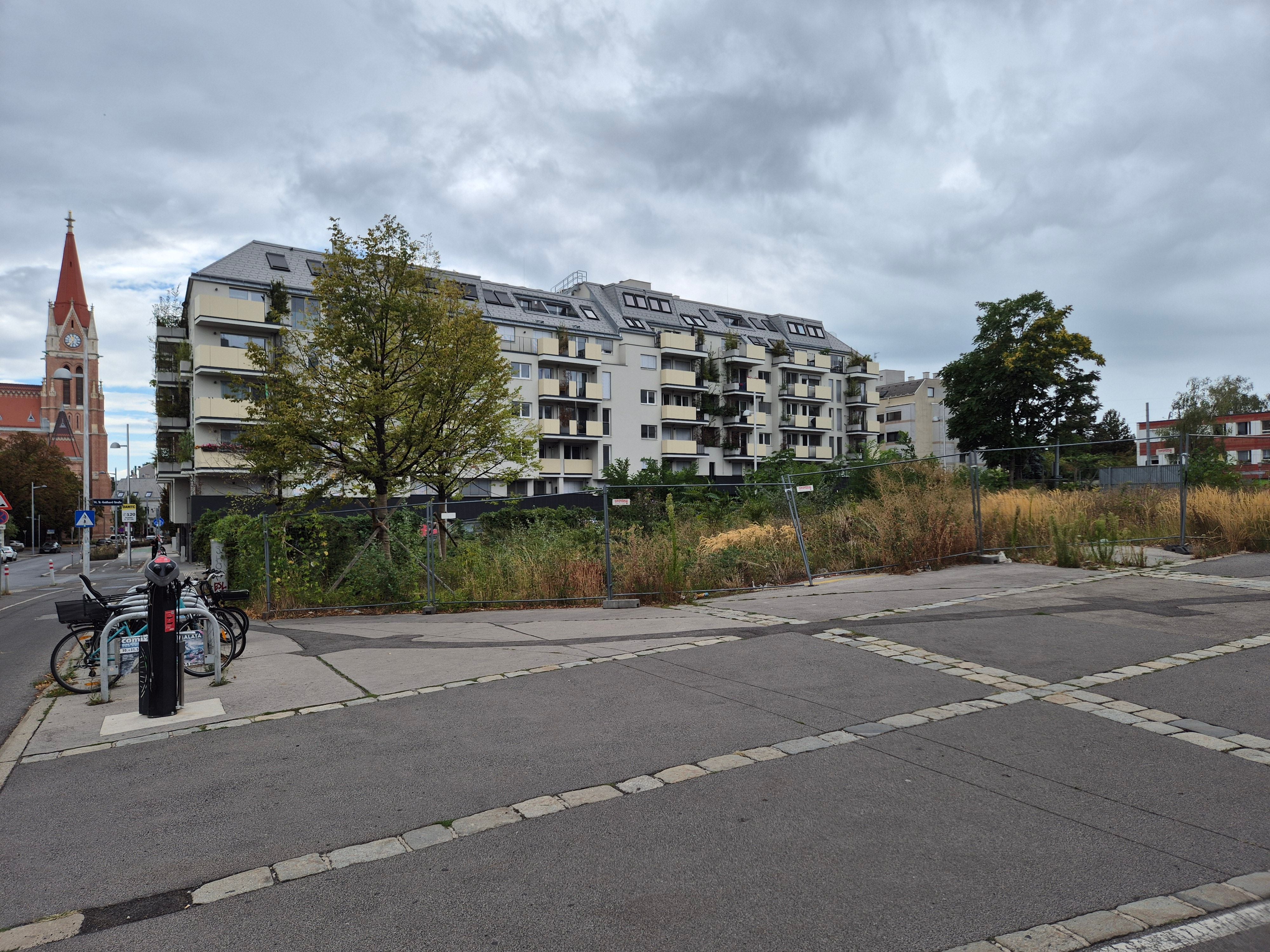 Wohnhaus mit Balkonen, davor ein leerstehendes, umzäuntes Grundstück und Fahrräder am Straßenrand.