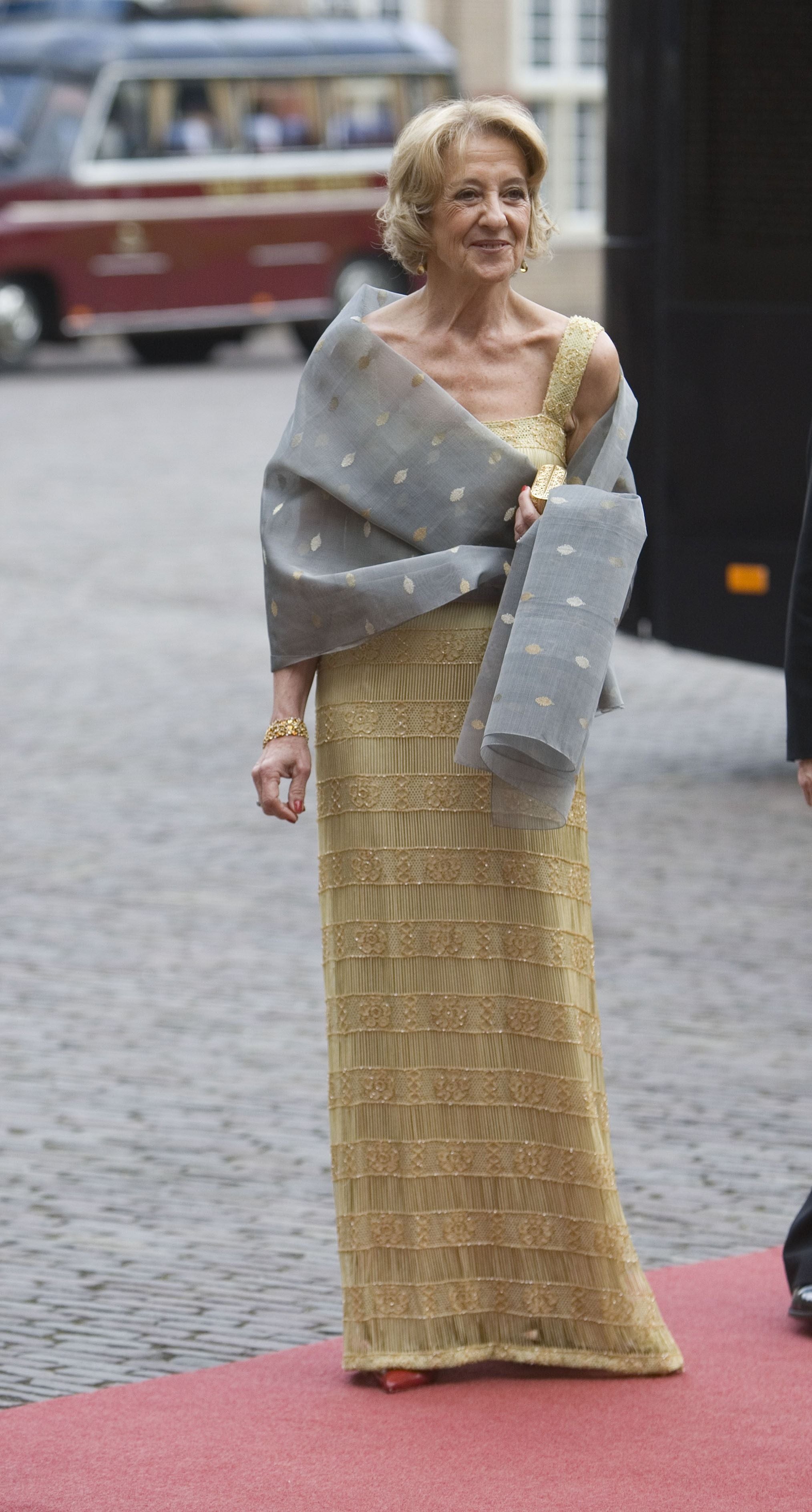 Elegante Frau in goldenem Kleid mit grauem Schal steht auf rotem Teppich im Freien.