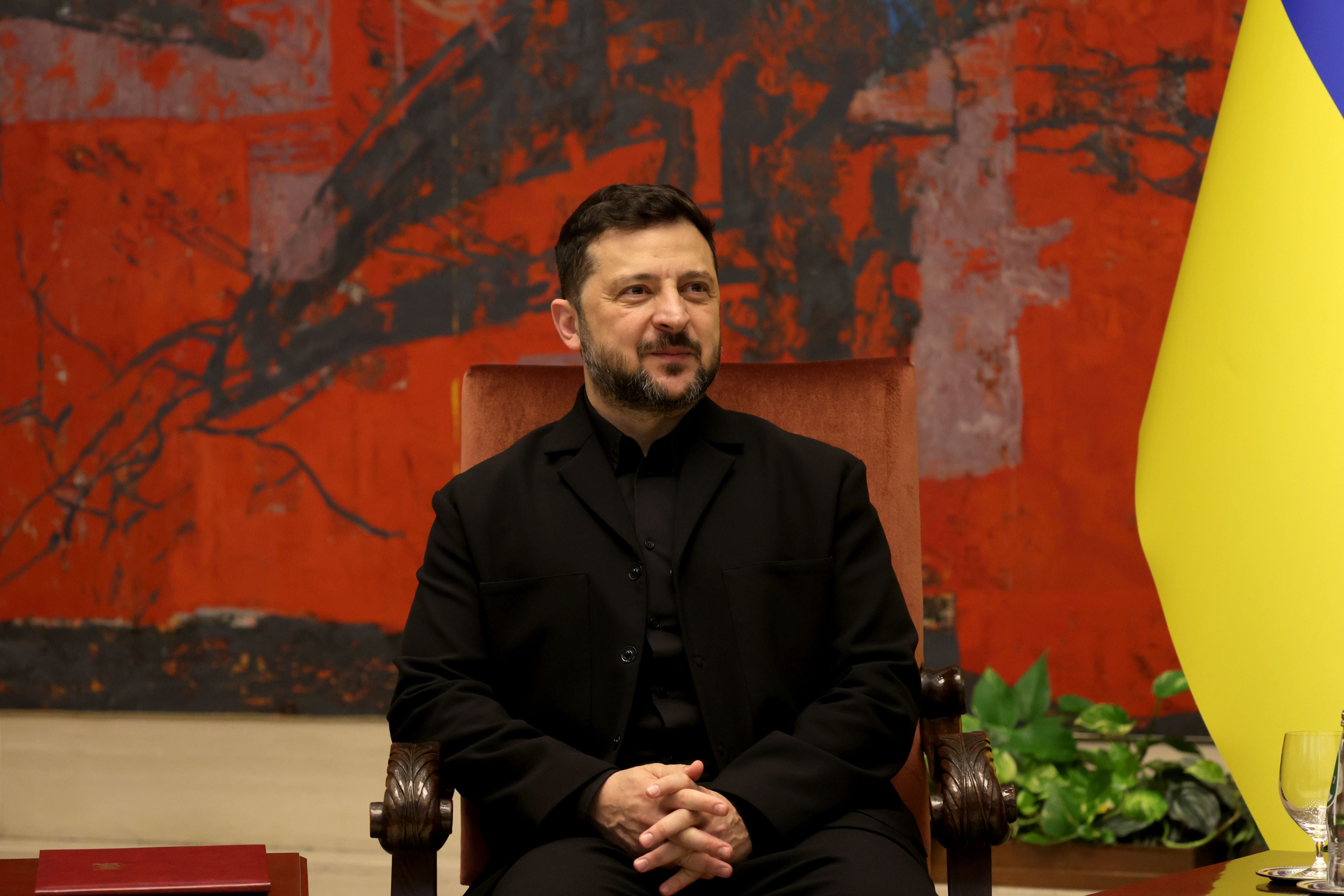 Ein Mann in schwarzer Kleidung sitzt vor einer roten Wand und einer ukrainischen Flagge.