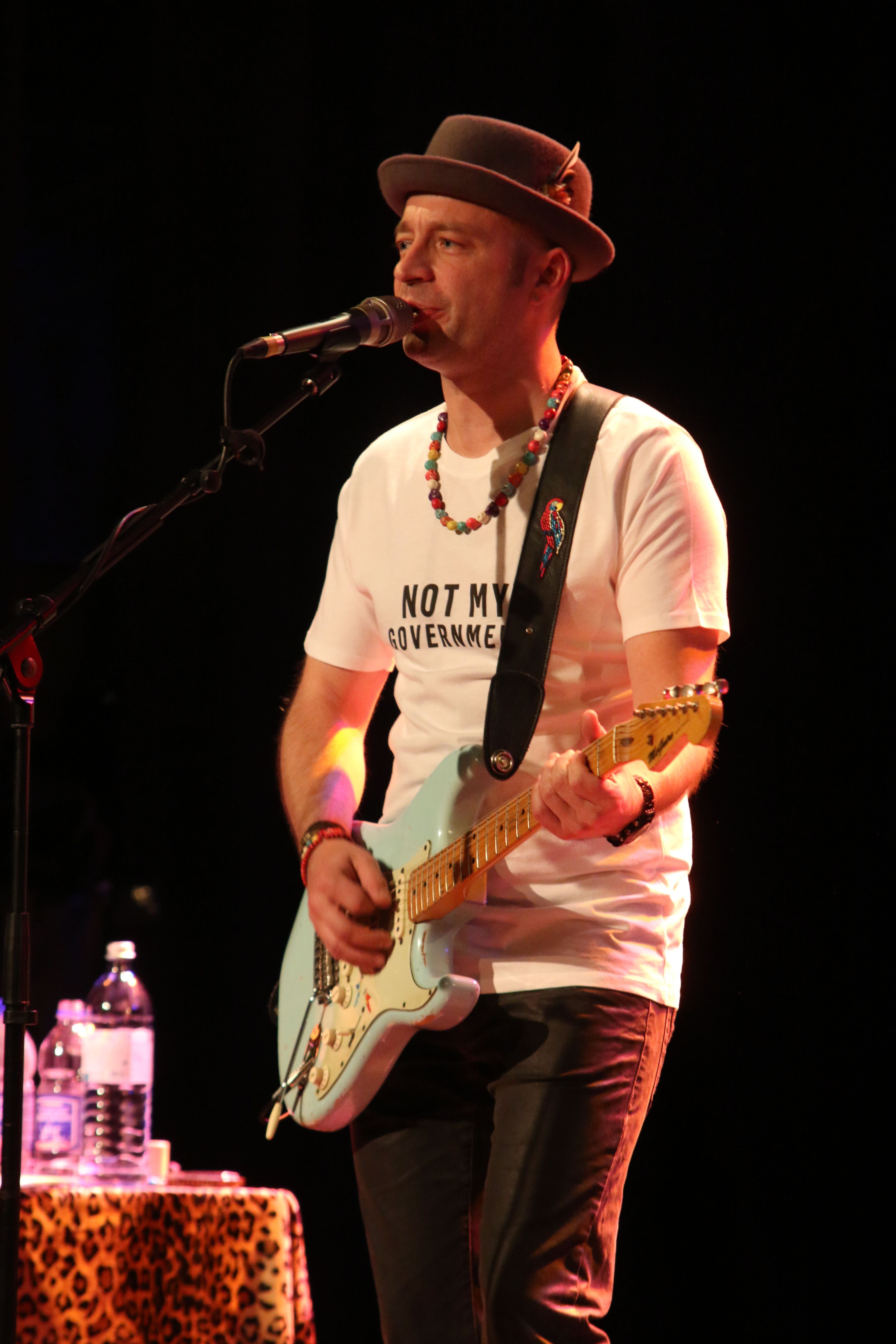 Mann mit Gitarre und Hut singt auf der Bühne, trägt ein "Not My Government"-T-Shirt.