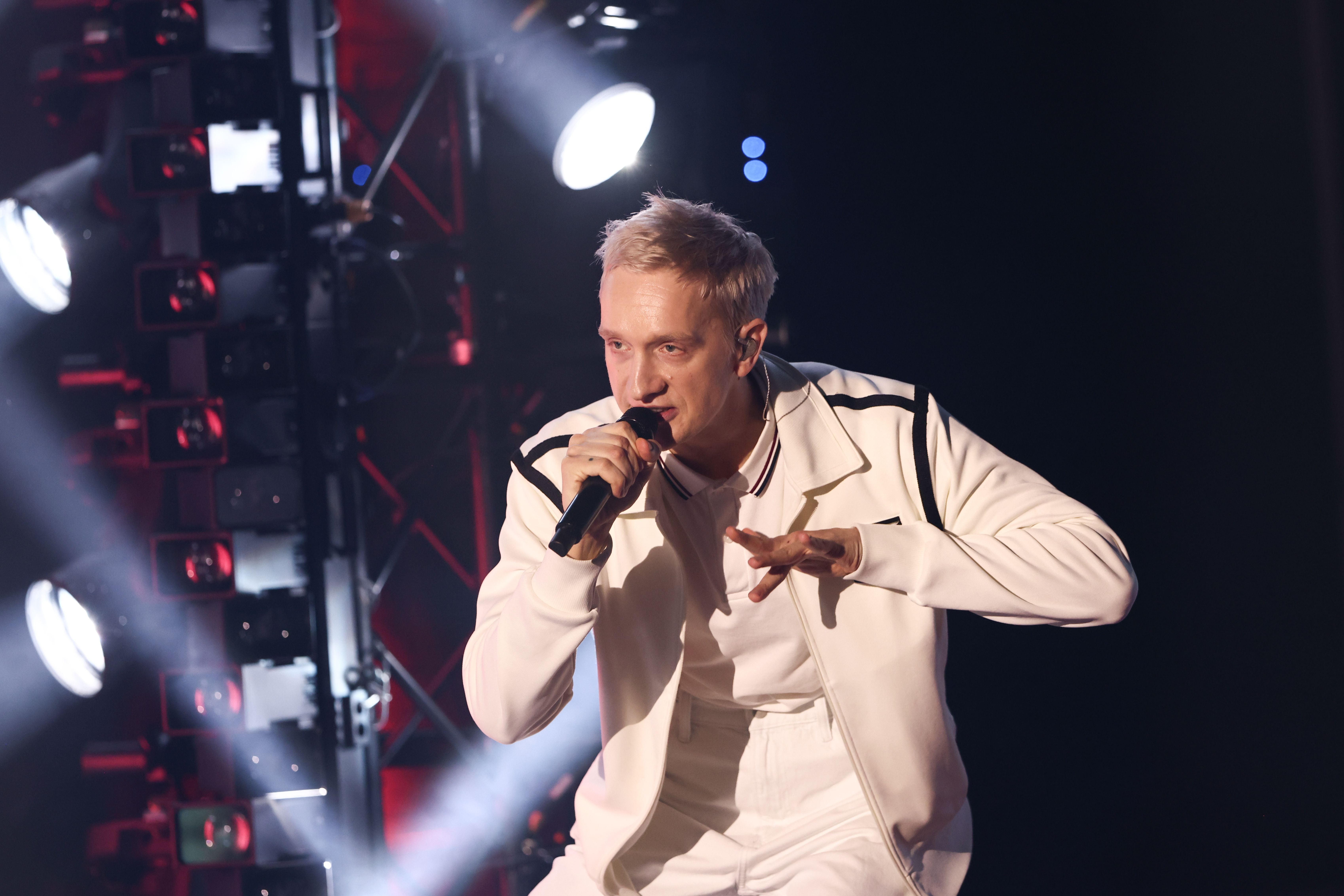 Ein Sänger in weißem Outfit tritt mit Mikrofon bei einem Konzert auf einer beleuchteten Bühne auf.
