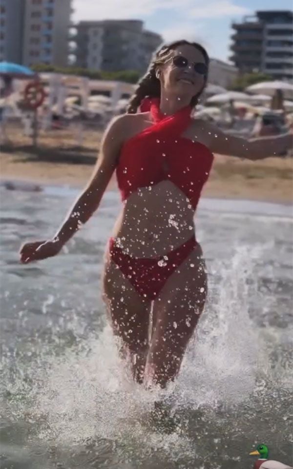 Frau in rotem Bikini läuft lachend durch flaches Wasser am Strand.