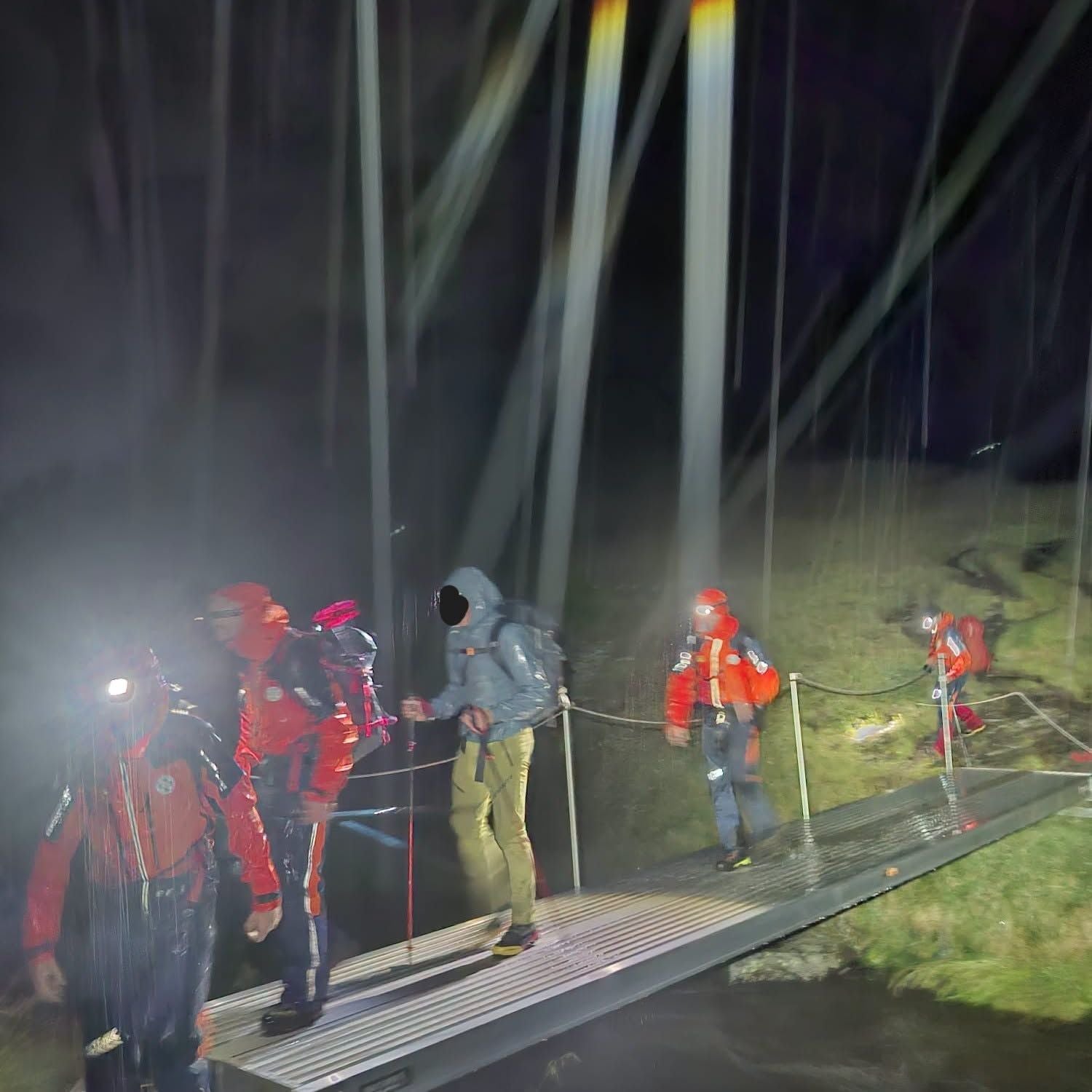Mehrere Rettungskräfte überqueren nachts bei Regen eine kleine Brücke im Gebirge.