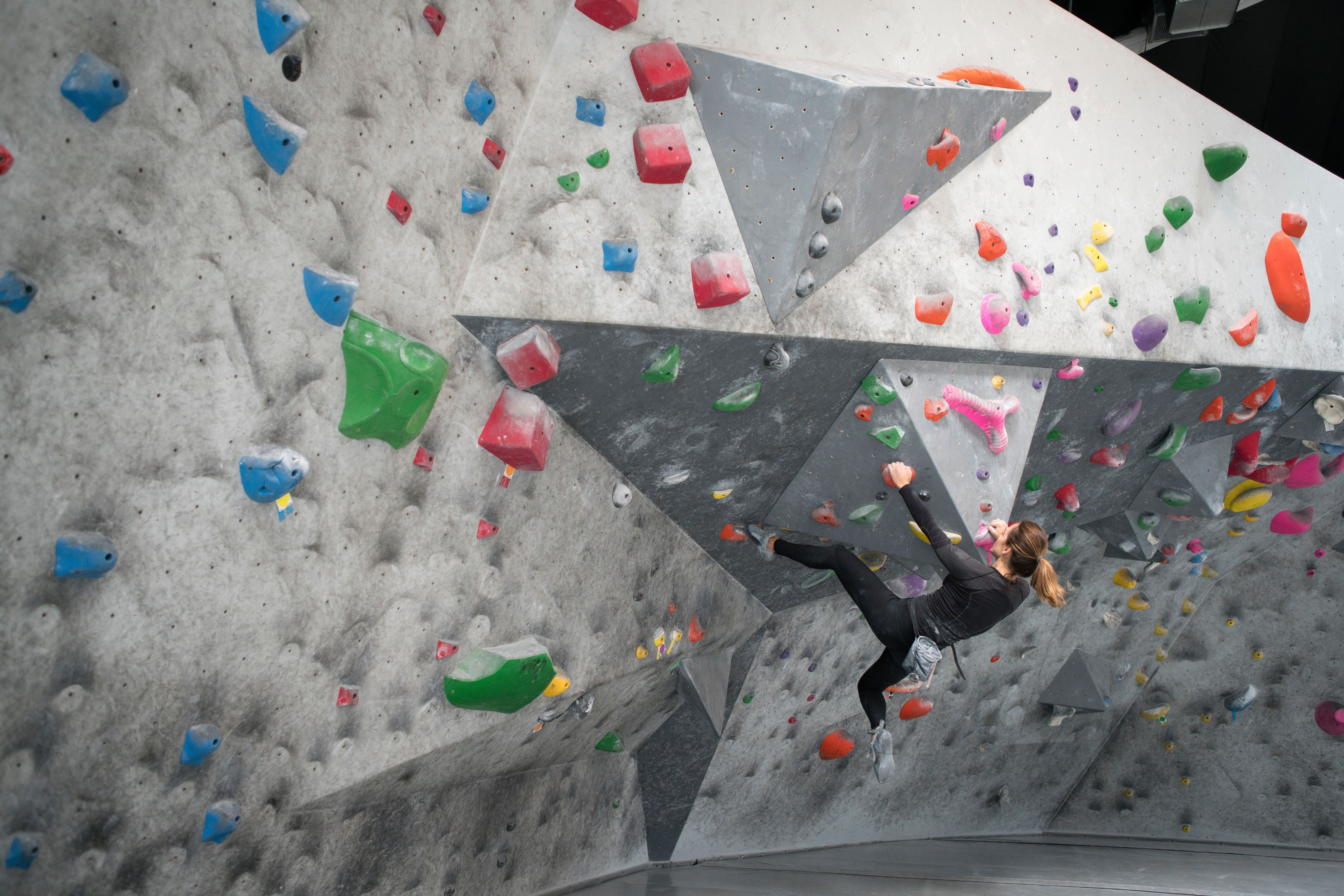 Frau klettert an einer Indoor-Boulderwand mit bunten Griffen und Henkeln.