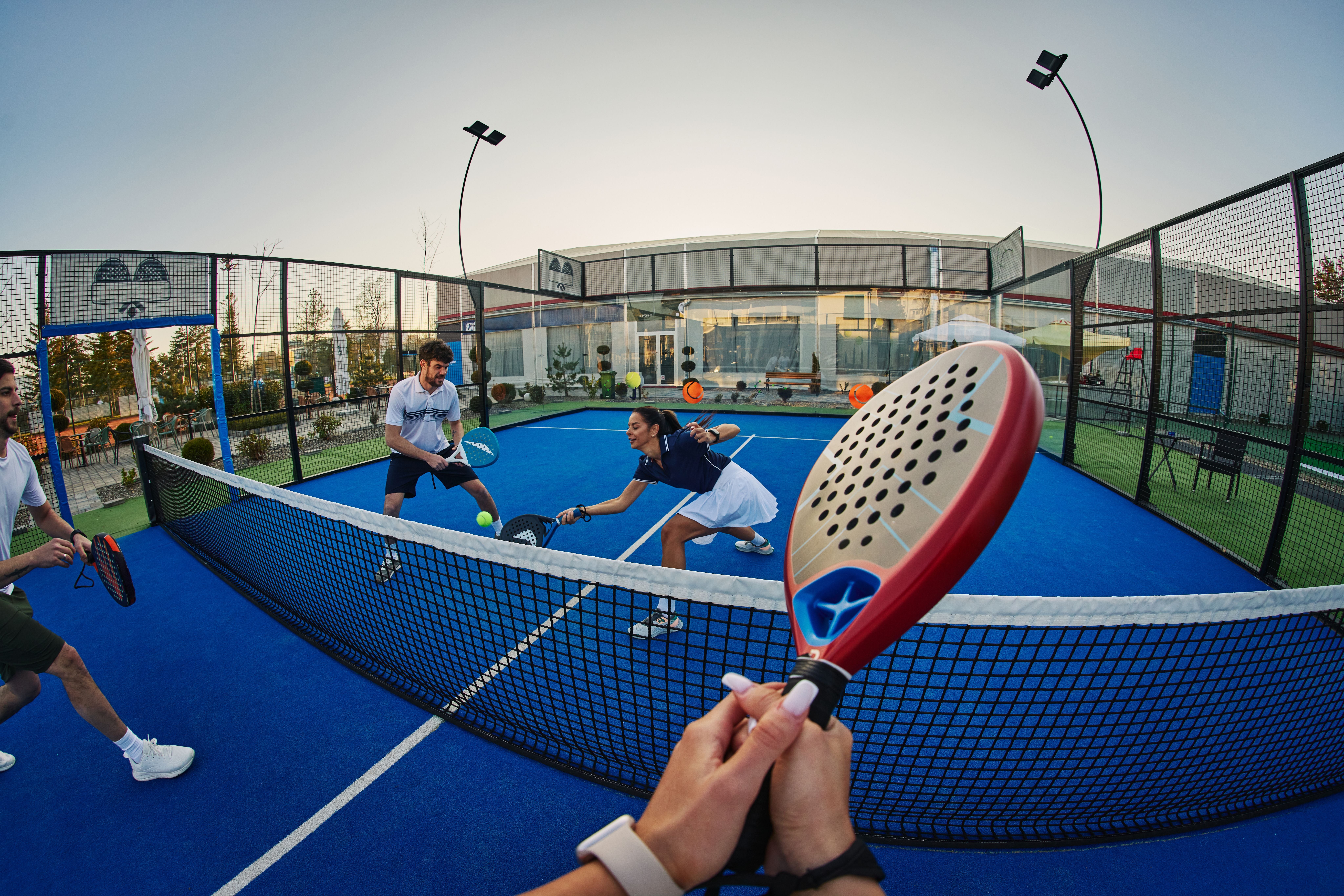 Freunde spielen Padel auf einem blauen Außenplatz, aufgenommen aus der Ich-Perspektive.