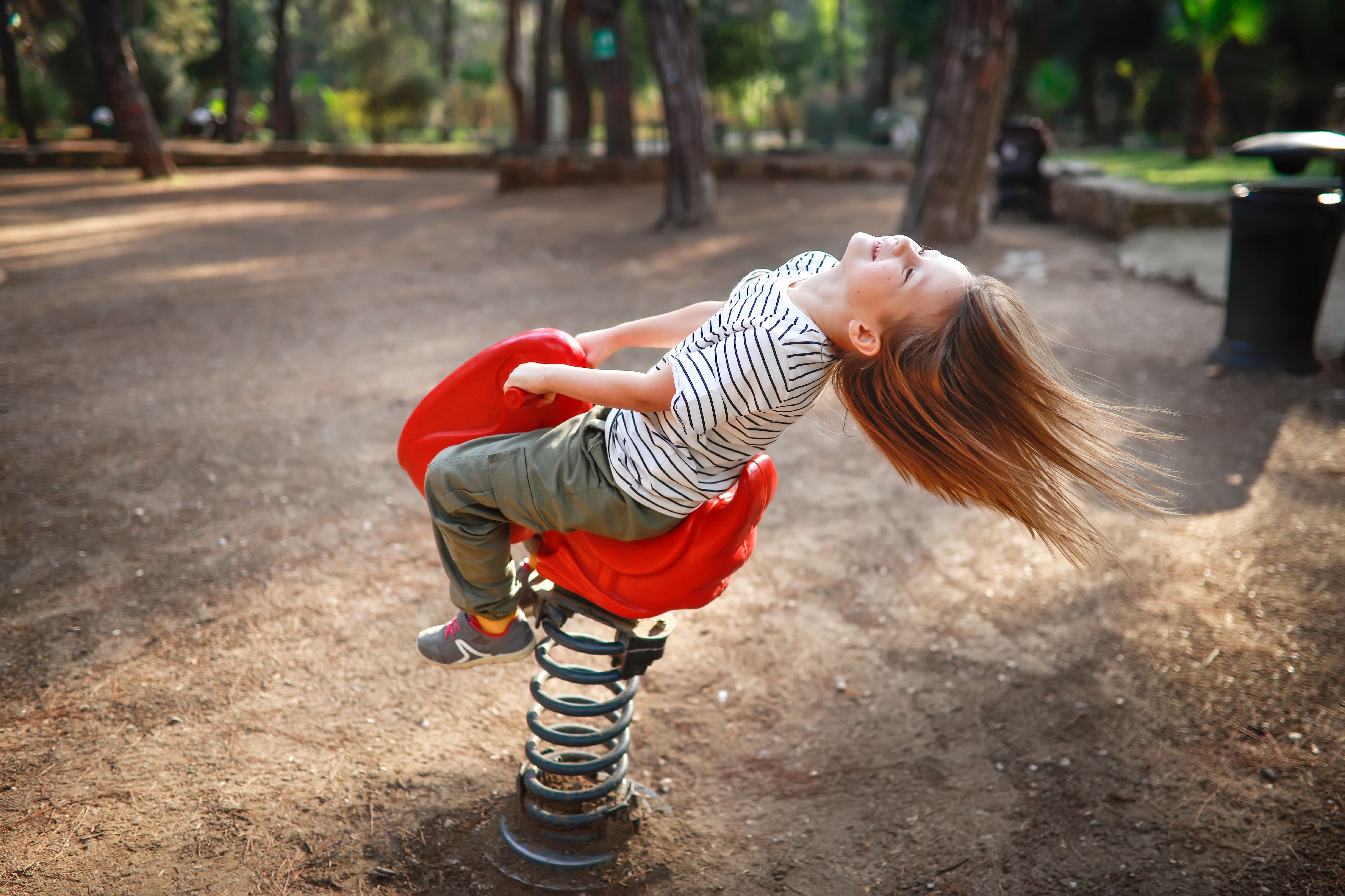 Kind lacht und schaukelt auf roter Federwippe im Parkspielplatz, Haare fliegen nach hinten.