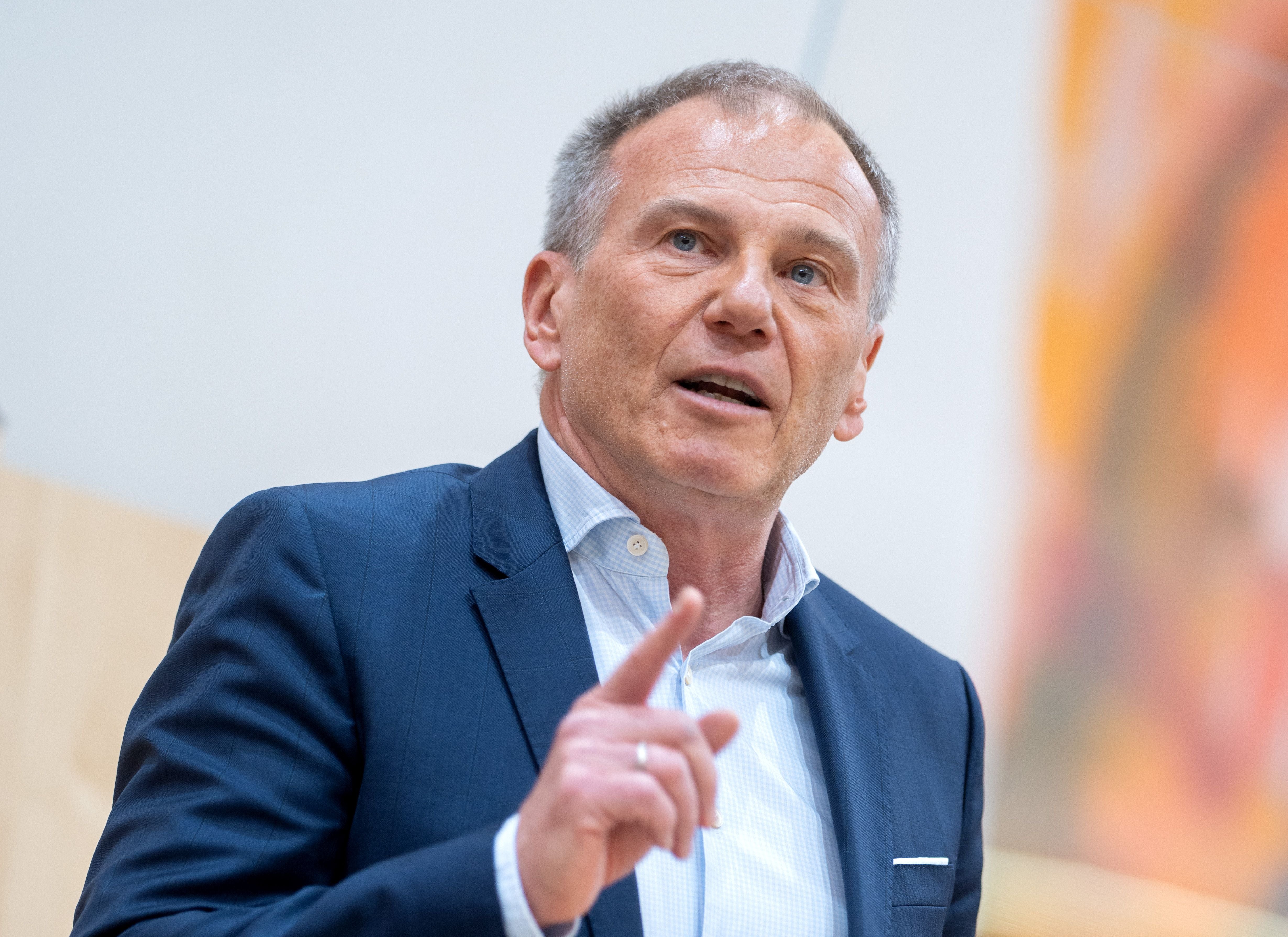 Ein Mann im Anzug spricht gestikulierend bei einer Veranstaltung mit hellem Hintergrund.