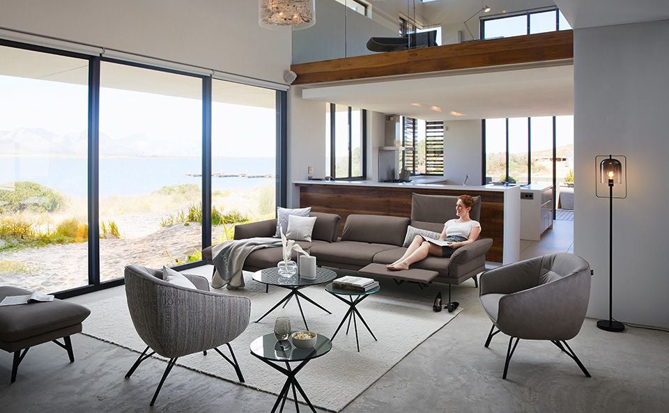 Modernes Wohnzimmer mit großen Fenstern und Blick auf einen See, Frau sitzt entspannt auf dem Sofa.