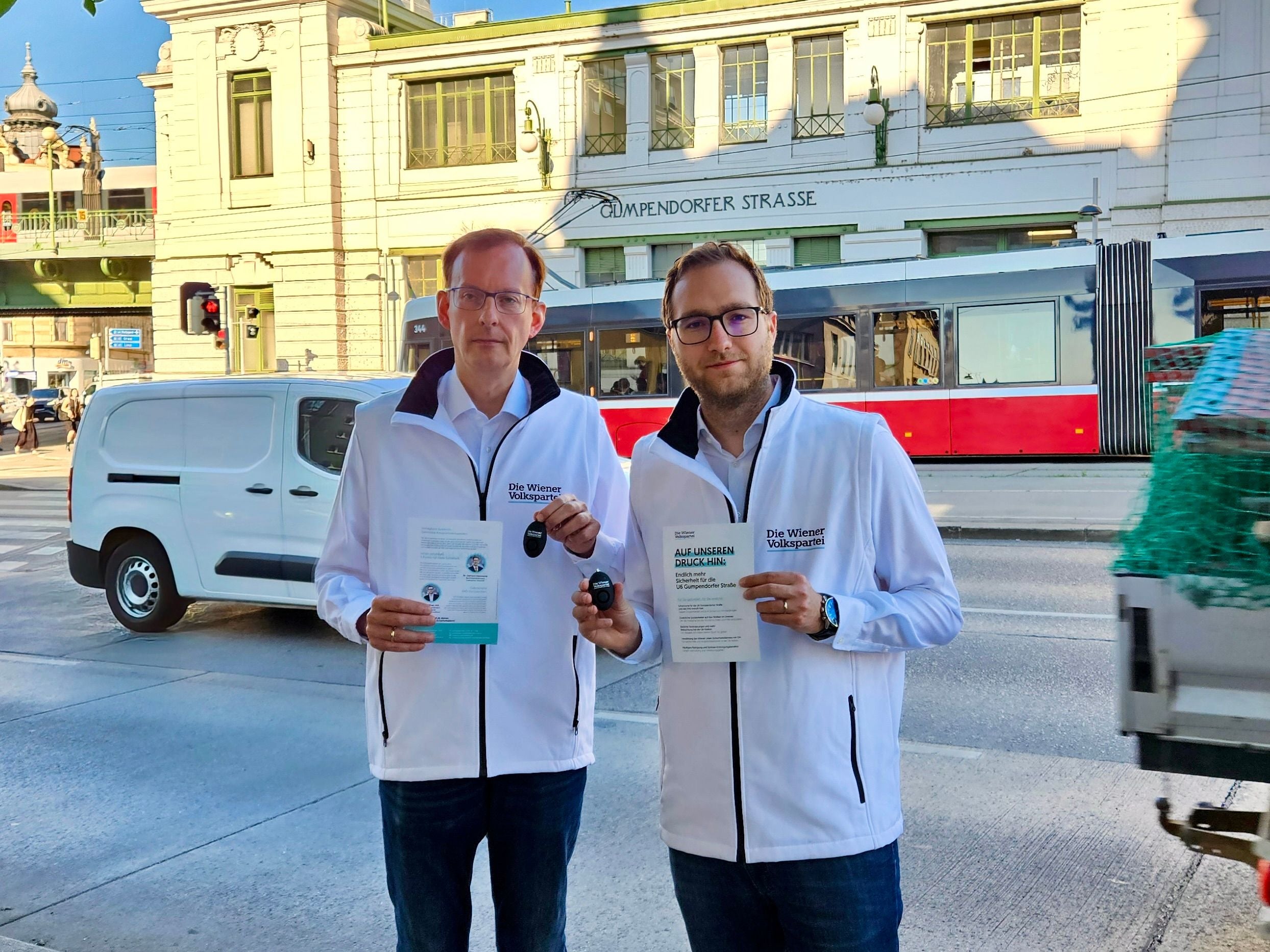 Zwei Männer in weißen Jacken halten Flyer auf einer Straße vor einer Straßenbahn.