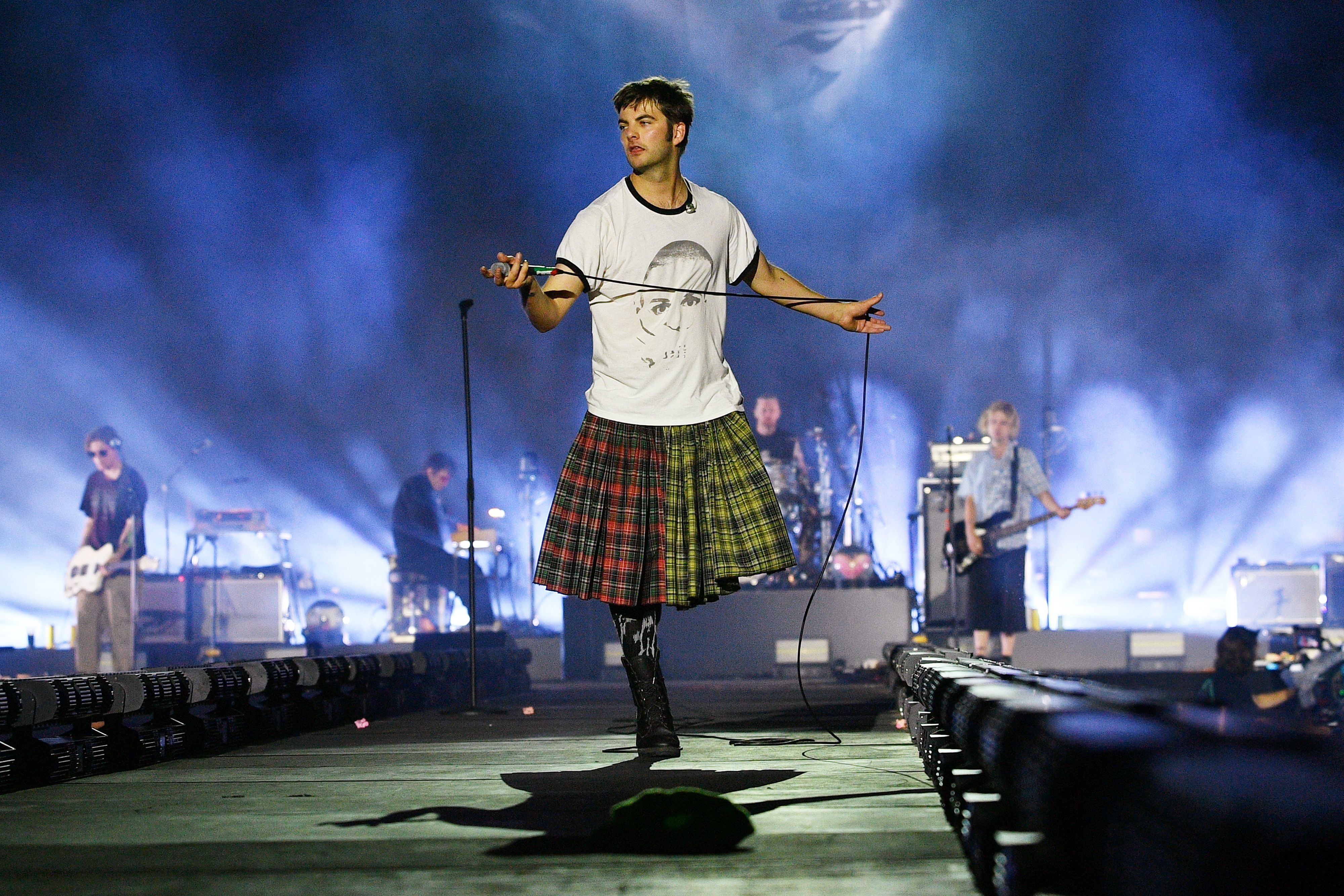 Sänger in kariertem Kilt und T-Shirt auf einer Konzertbühne mit Band im Hintergrund.