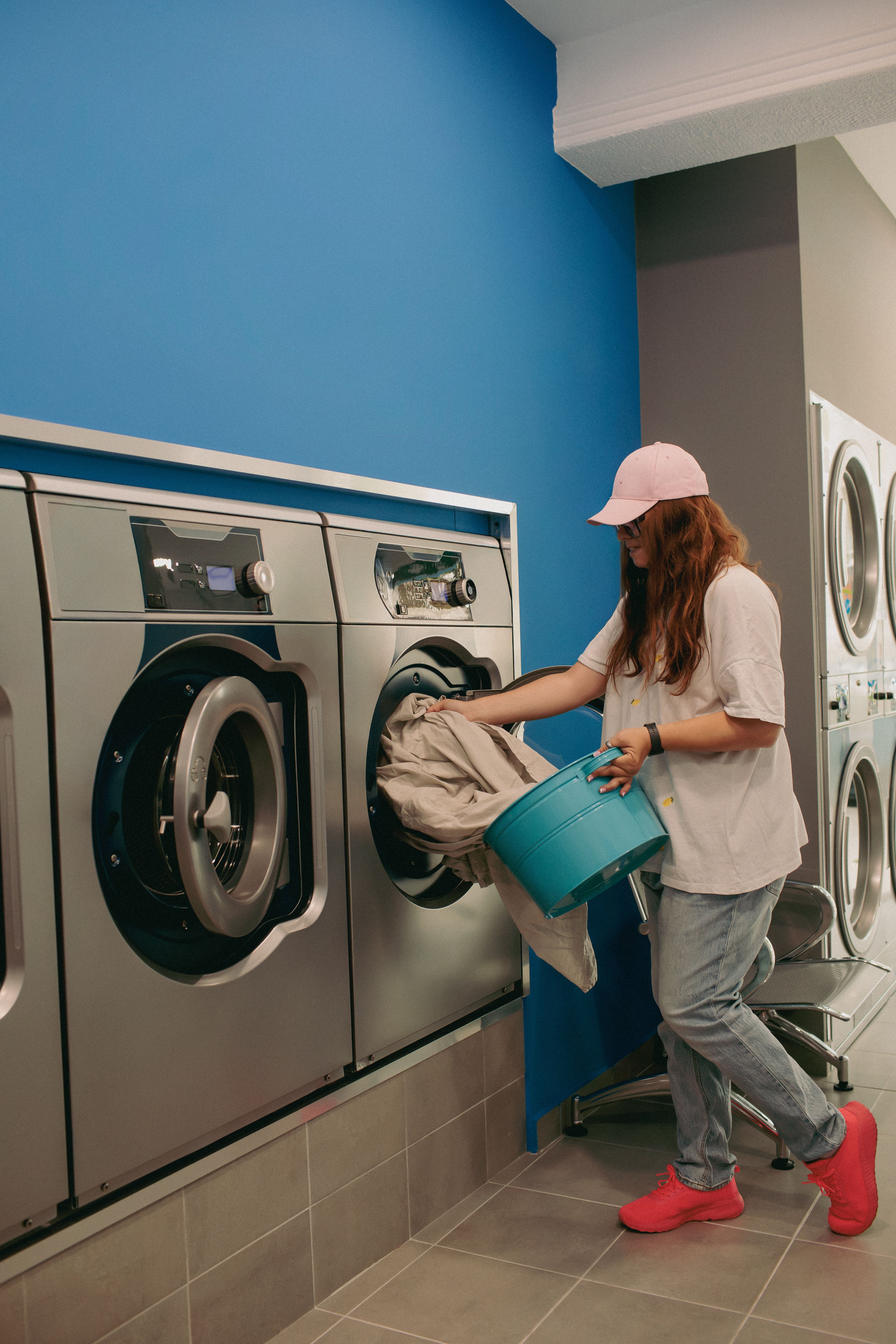Frau lädt Wäsche aus einem blauen Korb in eine moderne Waschmaschine im Waschsalon.