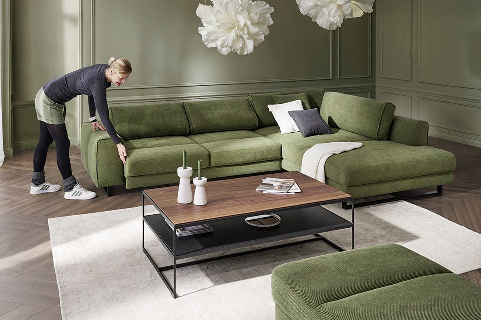 Frau richtet grünes Ecksofa im Wohnzimmer mit Holz-Couchtisch und hellem Teppich aus.