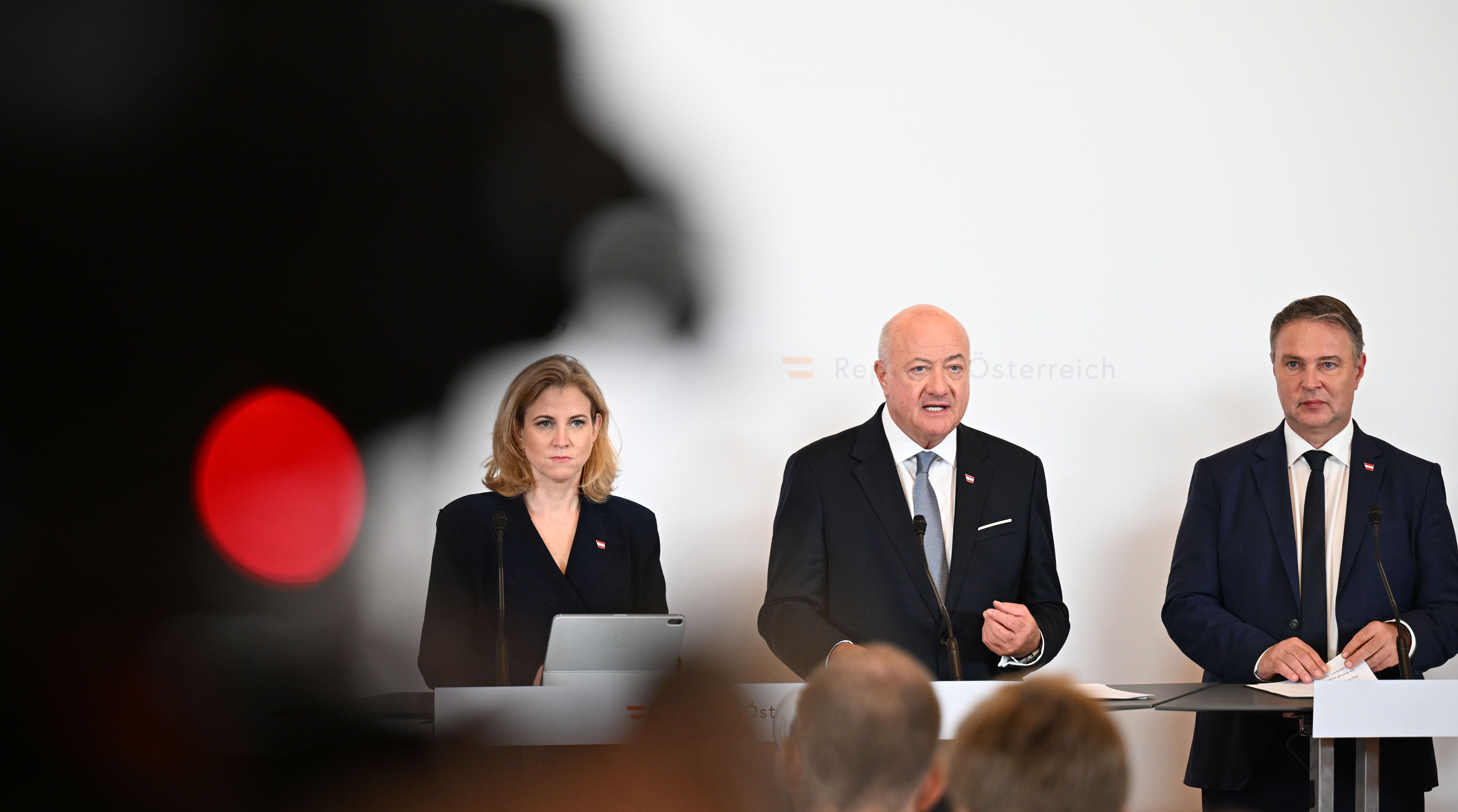 Drei Politiker stehen bei einer Pressekonferenz mit Mikrofonen und Notizen vor weißem Hintergrund.