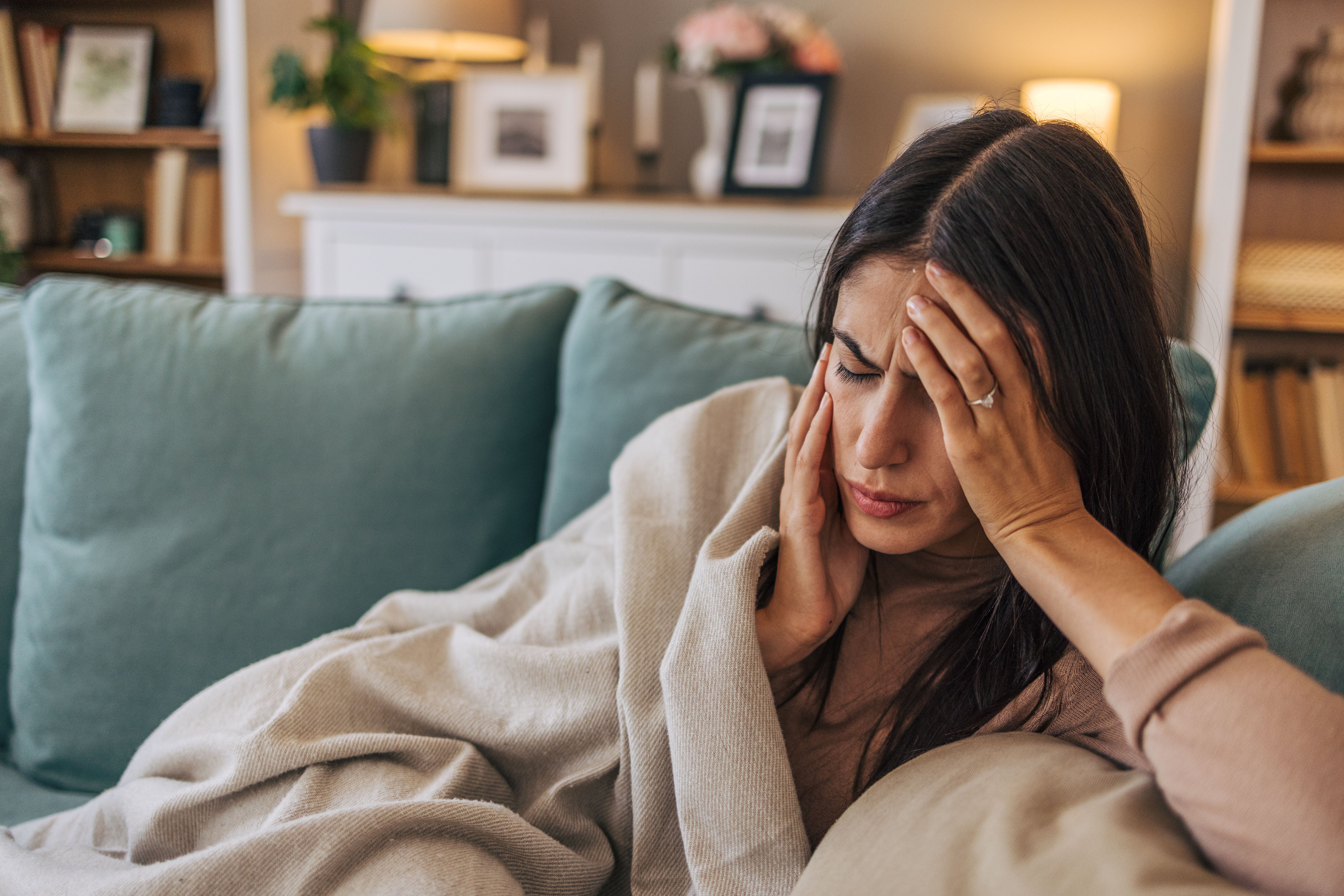 Junge Frau liegt krank auf dem Sofa und hält sich den Kopf vor Schmerzen.