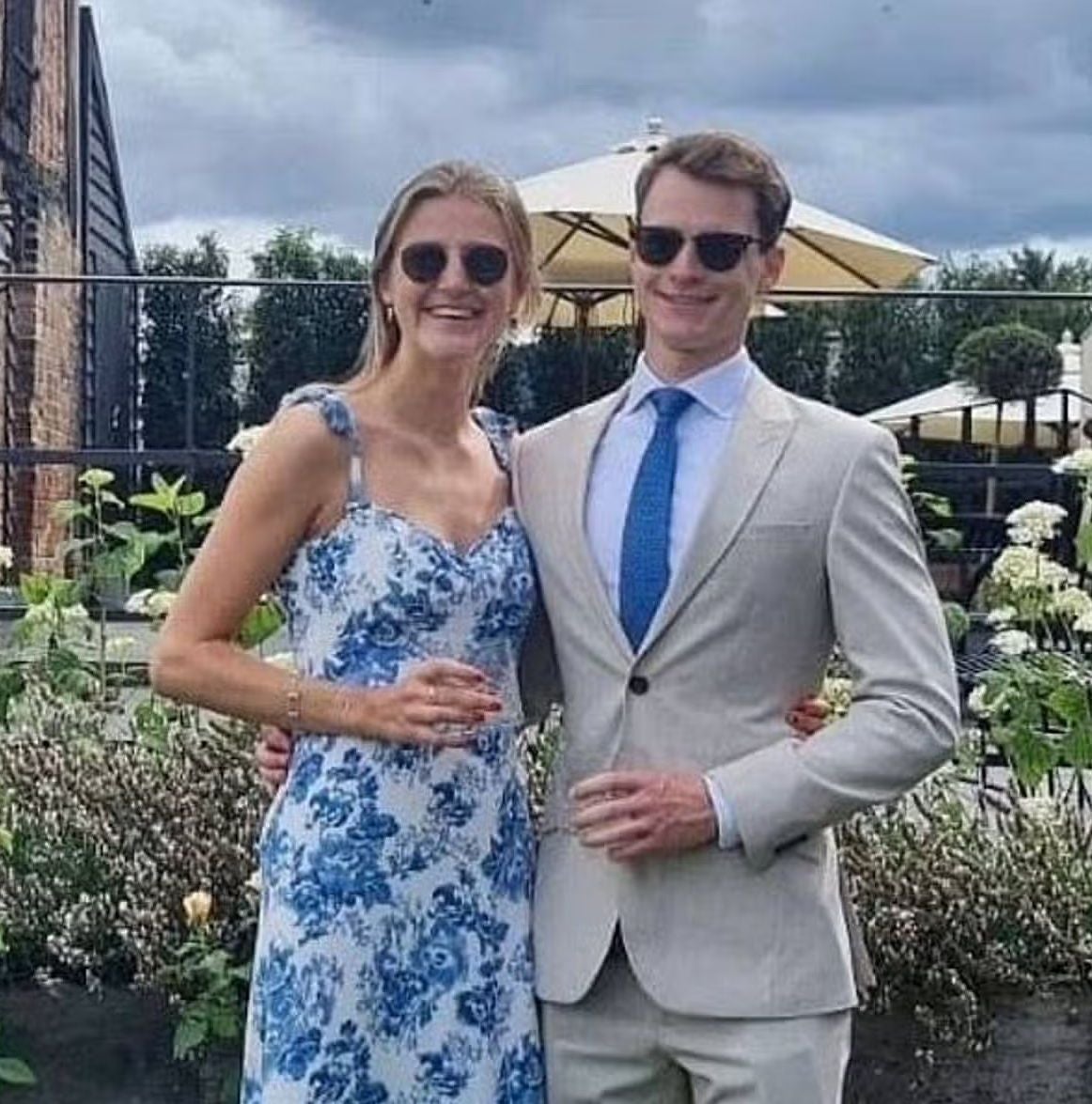 Ein Paar in eleganter Kleidung posiert draußen, beide tragen Sonnenbrillen und lächeln.