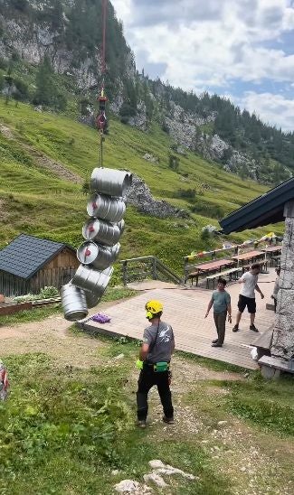 Mehrere Bierfässer hängen an einem Kran; Menschen beobachten die Anlieferung im Gebirge.