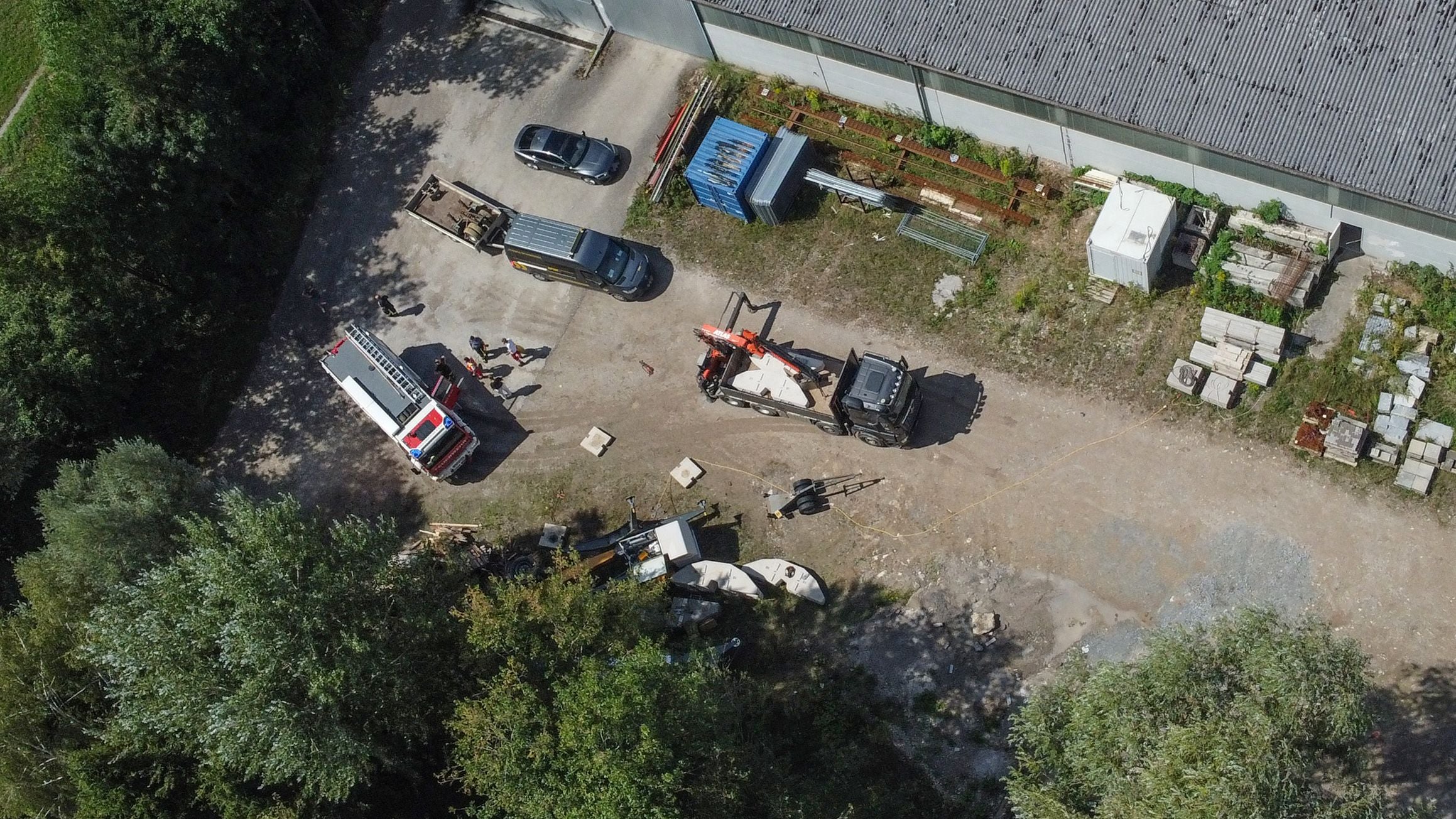 Luftaufnahme eines Firmengeländes mit Kranfahrzeug, Autos, Feuerwehr und einigen Personen.