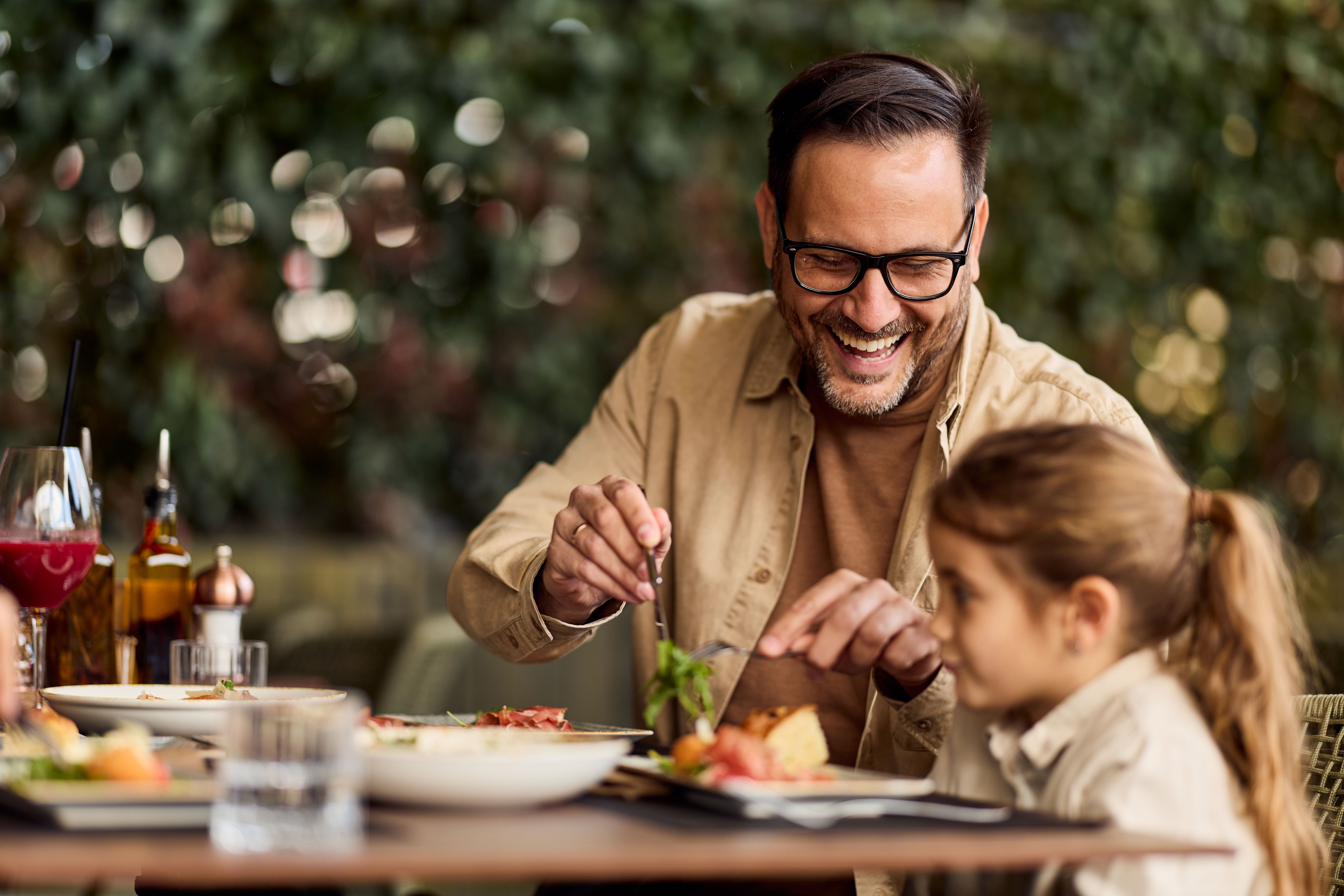 Vater und Tochter genießen lachend ein gemeinsames Mittagessen auf einer Restaurant-Terrasse.
