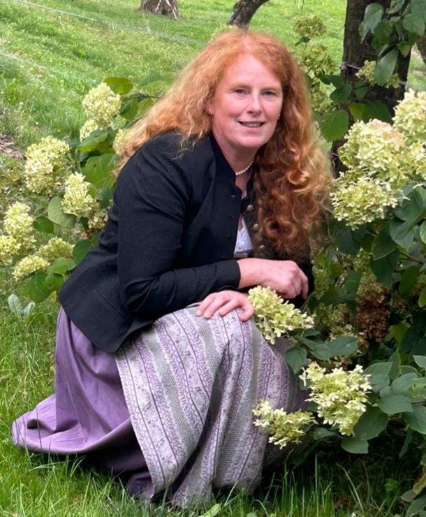 Frau mit langen roten Haaren kniet im Garten neben weißen Blüten und lächelt in die Kamera.