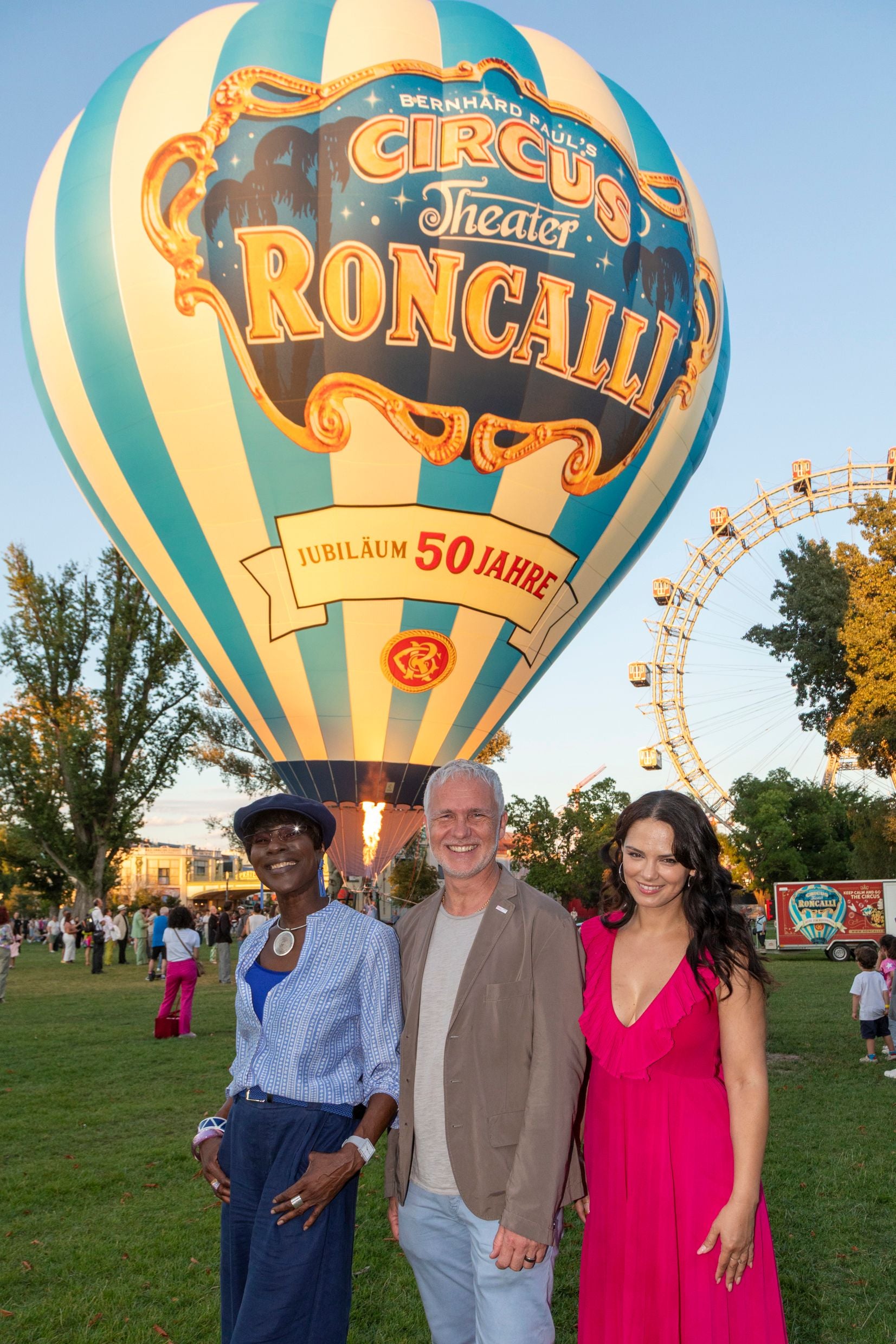Drei Menschen posieren vor einem Roncalli-Heißluftballon bei einer Präsentation in Wien.