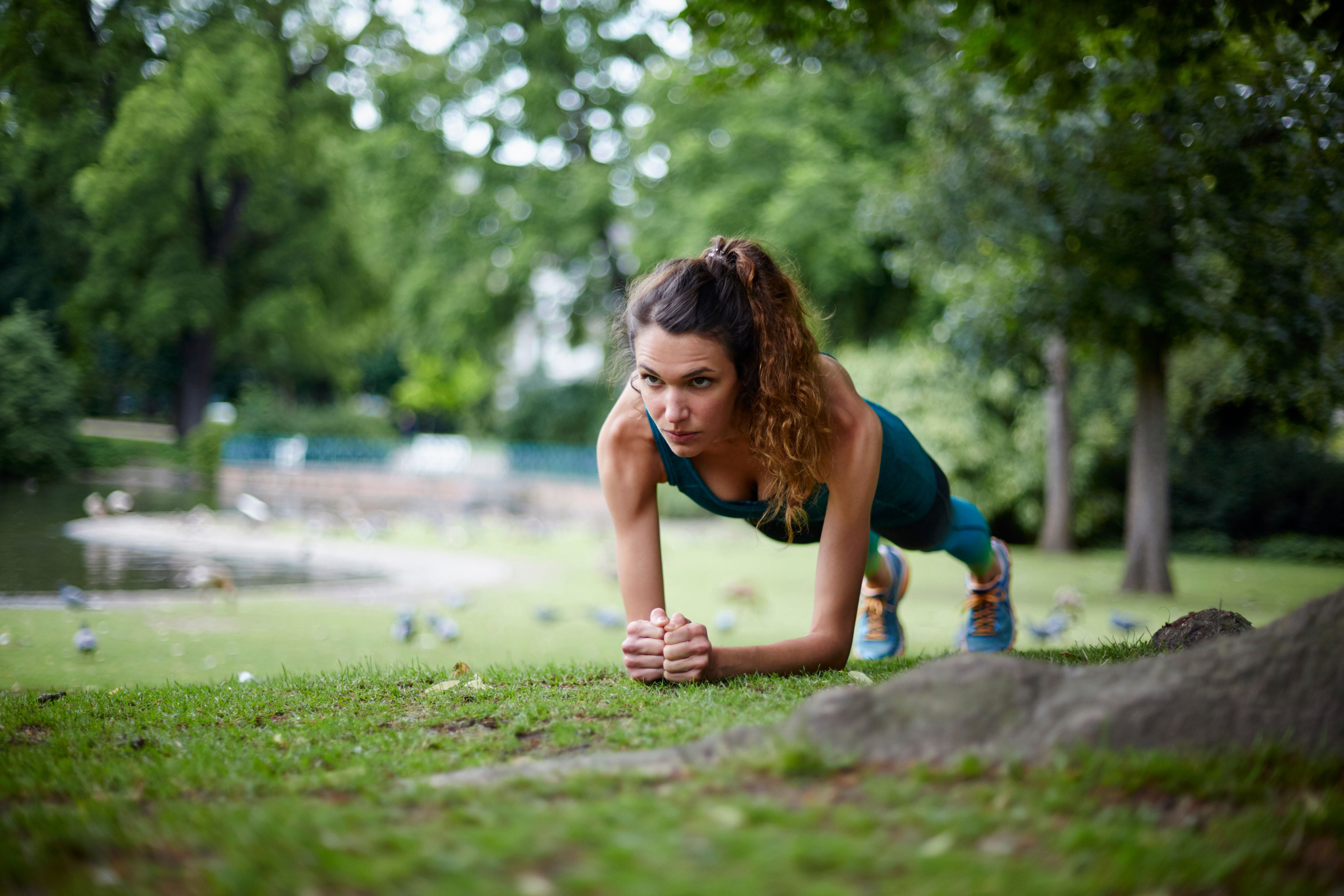 Frau macht Plank-Übung auf einer Wiese im Park, Bäume im Hintergrund.