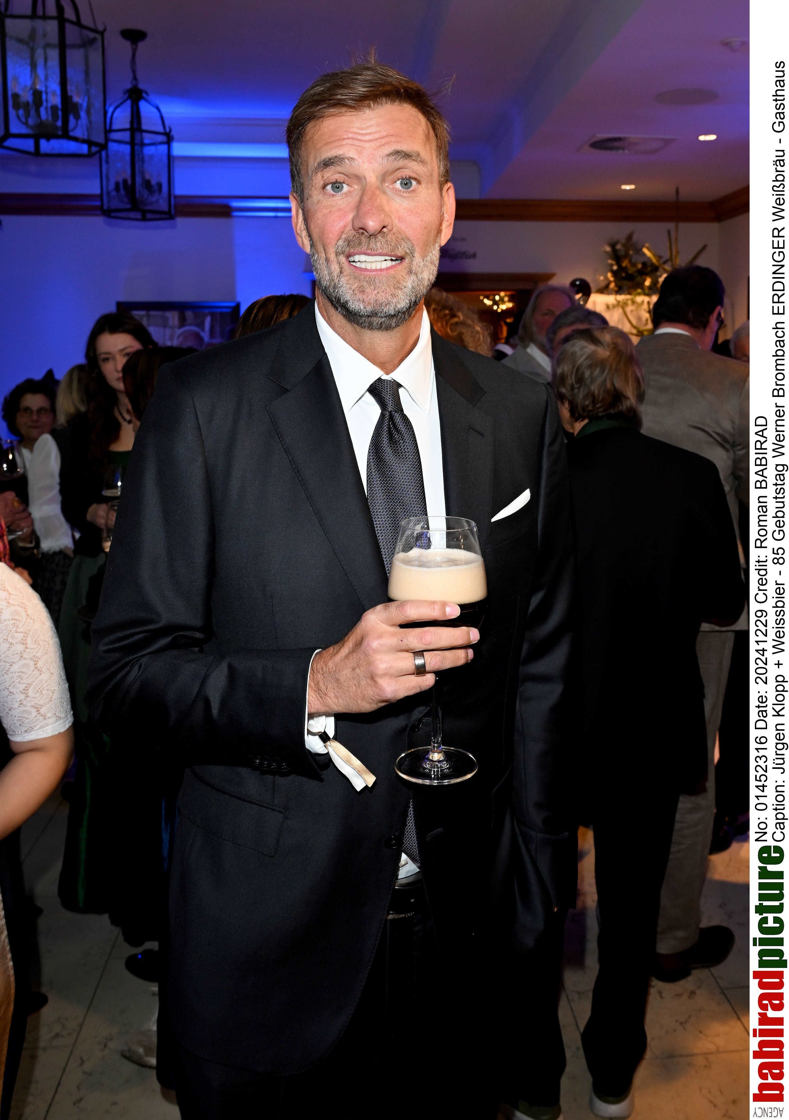 Mann im Anzug hält ein Glas Weißbier bei einer Feier mit mehreren Gästen im Hintergrund.