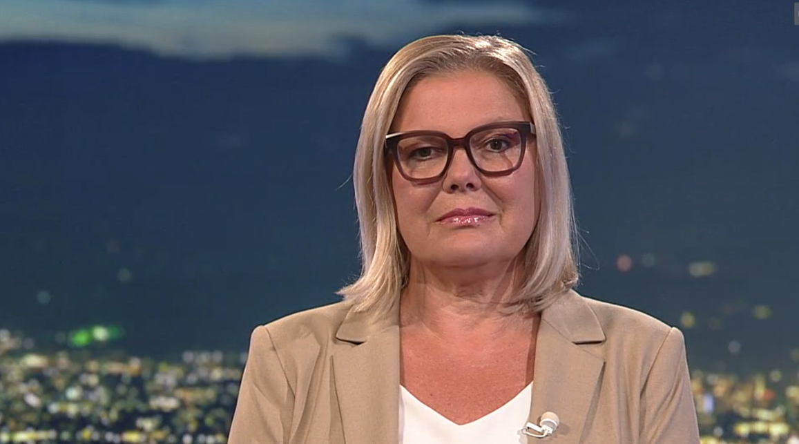 Frau mit blonden Haaren, Brille und beigem Blazer vor verschwommenem Hintergrund.