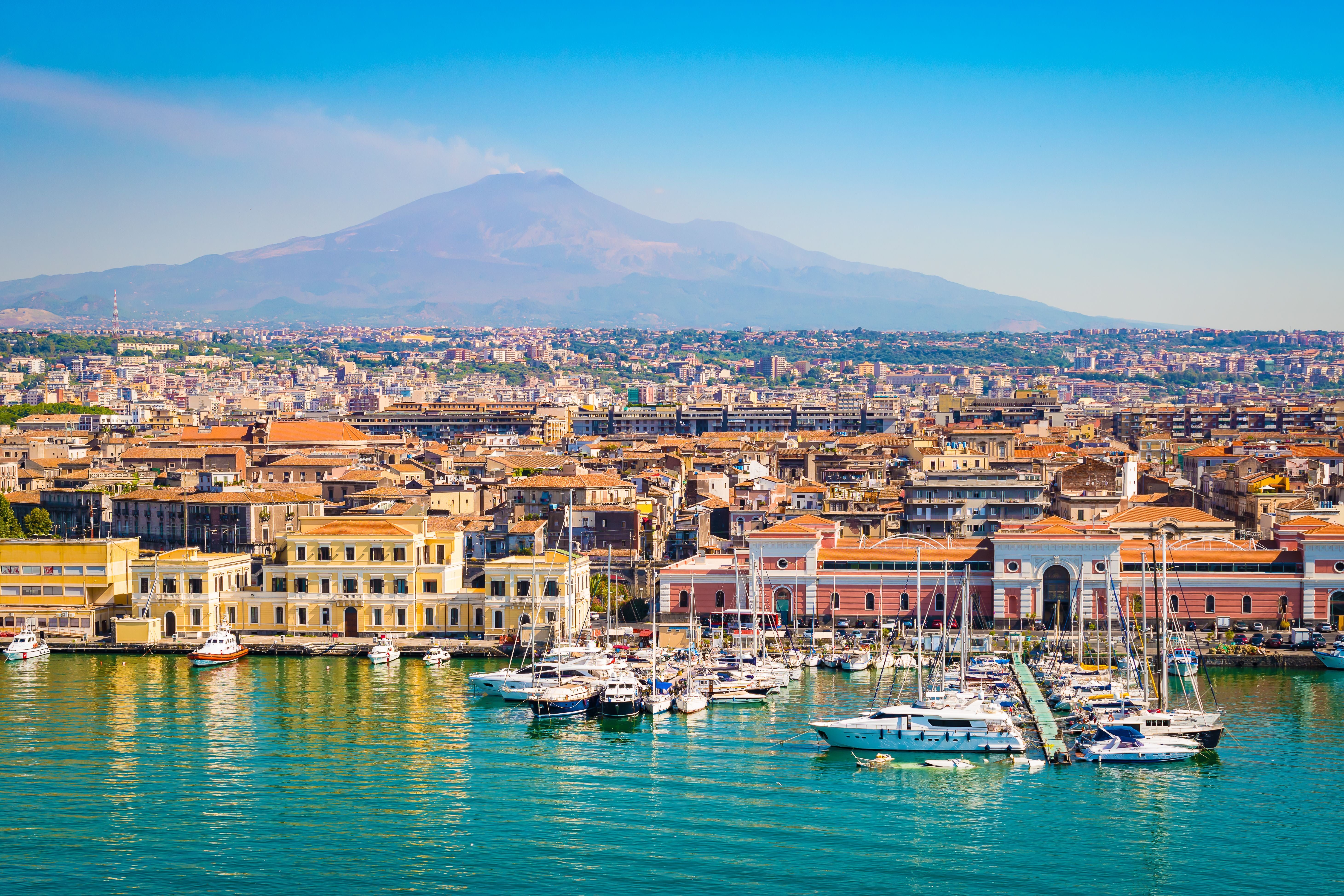 Hafen von Catania mit Yachten, dahinter Stadt und rauchender Vulkan Ätna im Hintergrund.